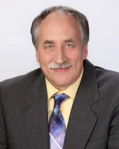 A smiling man with gray hair and a mustache wearing a gray suit, yellow shirt, and blue tie, against a plain white background.