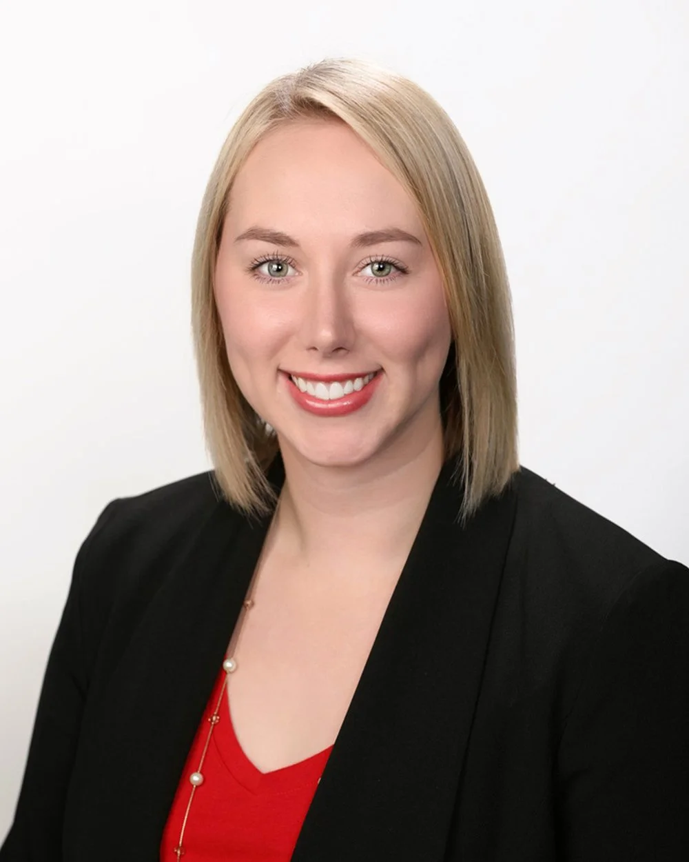 A woman with blonde hair, wearing a black blazer and red top, smiling against a white background.