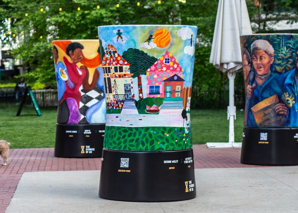Three decorated cylindrical sculptures with colorful artwork, set outdoors on a paved area with greenery and trees in the background. Each sculpture features unique, vibrant illustrations, and stands on black bases with QR codes and text.