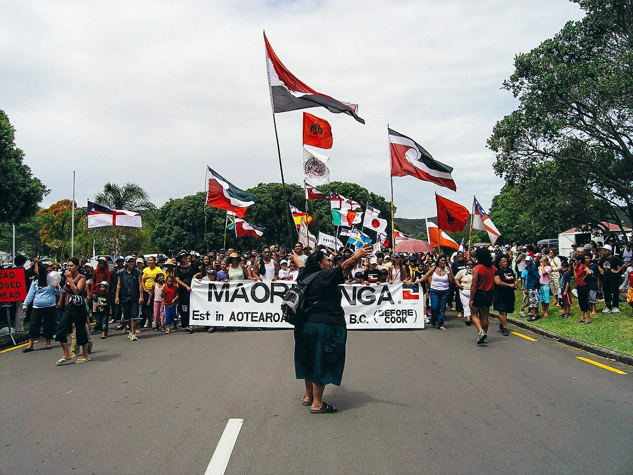 Protest march with people holding flags and a banner, on a road lined with trees, under overcast skies.