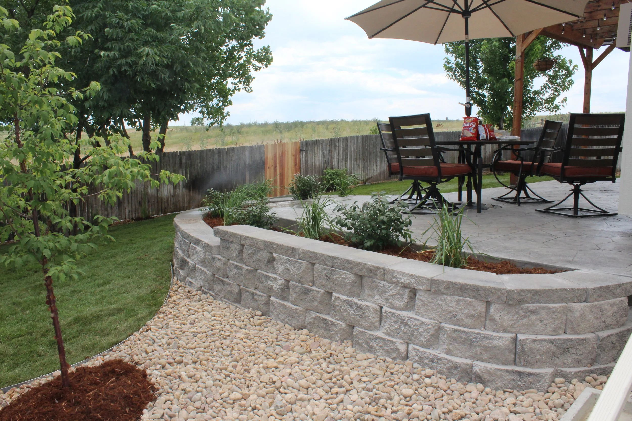 An outdoor patio with a stone retaining wall, a table with four chairs, a large umbrella, and trees and a grassy field in the background.
