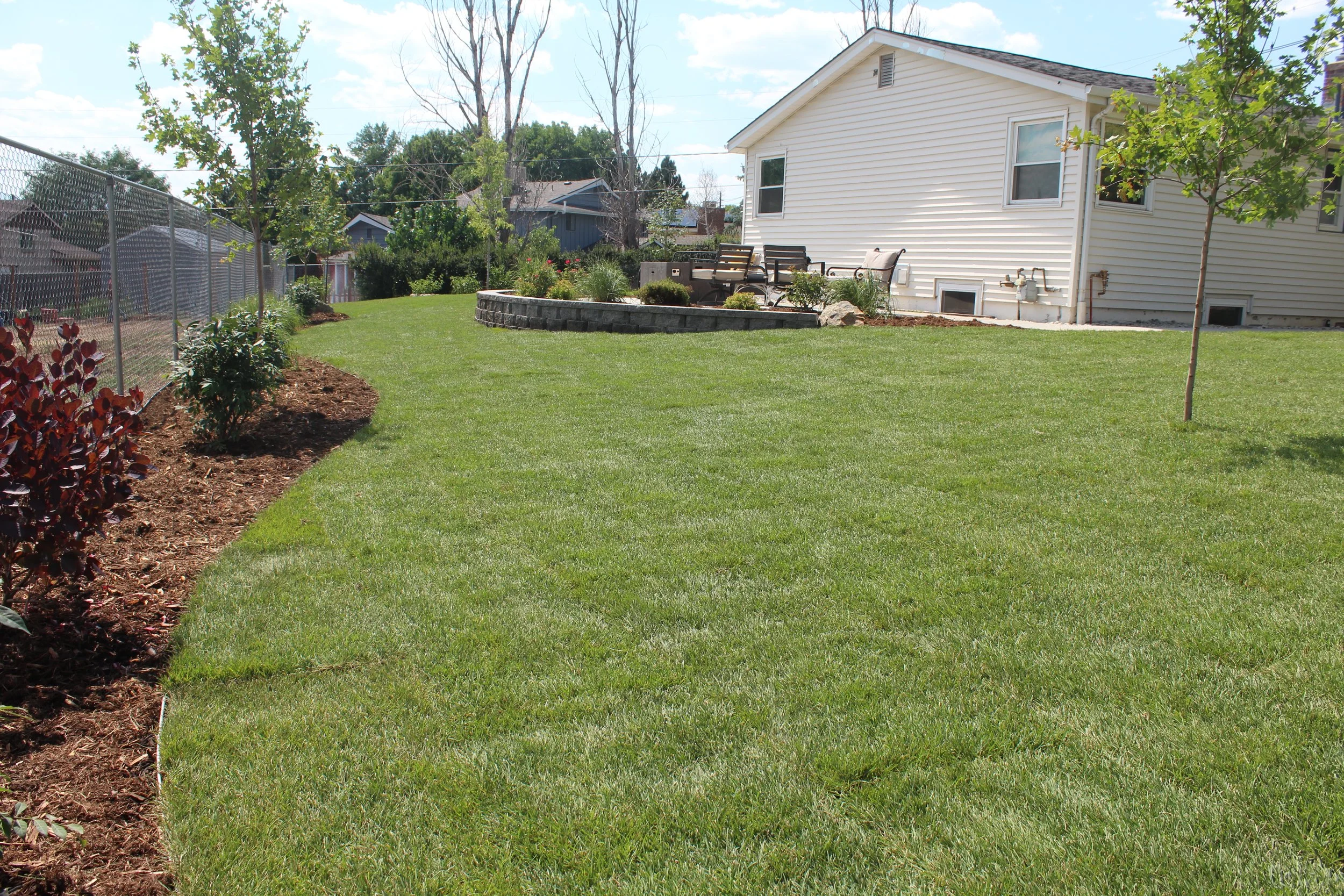A backyard lawn with a white house in the background, small trees, a landscaped flower bed with a curved stone border, outdoor furniture, and a chain-link fence on the left side.