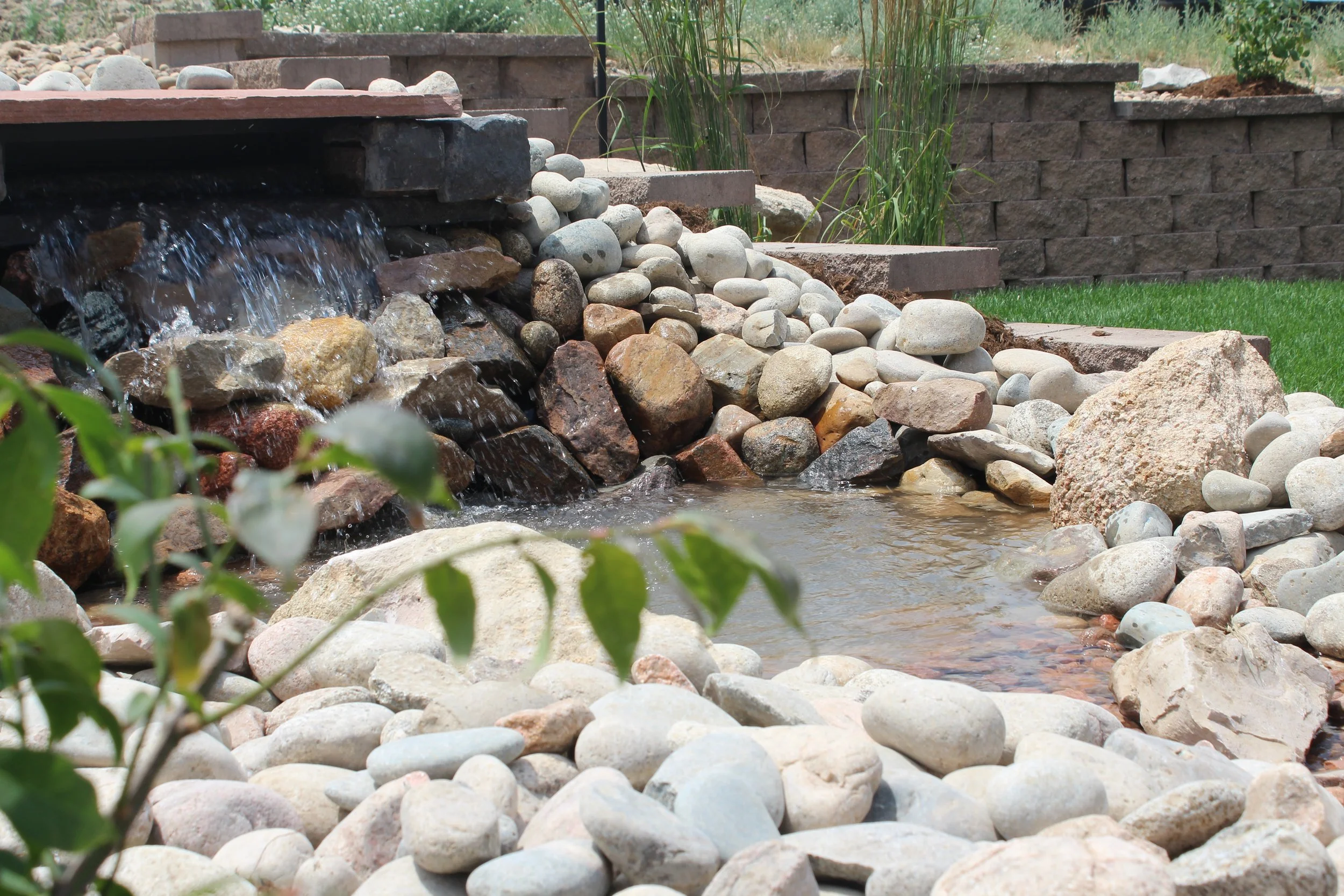 Large water fall with pond and cobble with retaining wall and grasses in the background