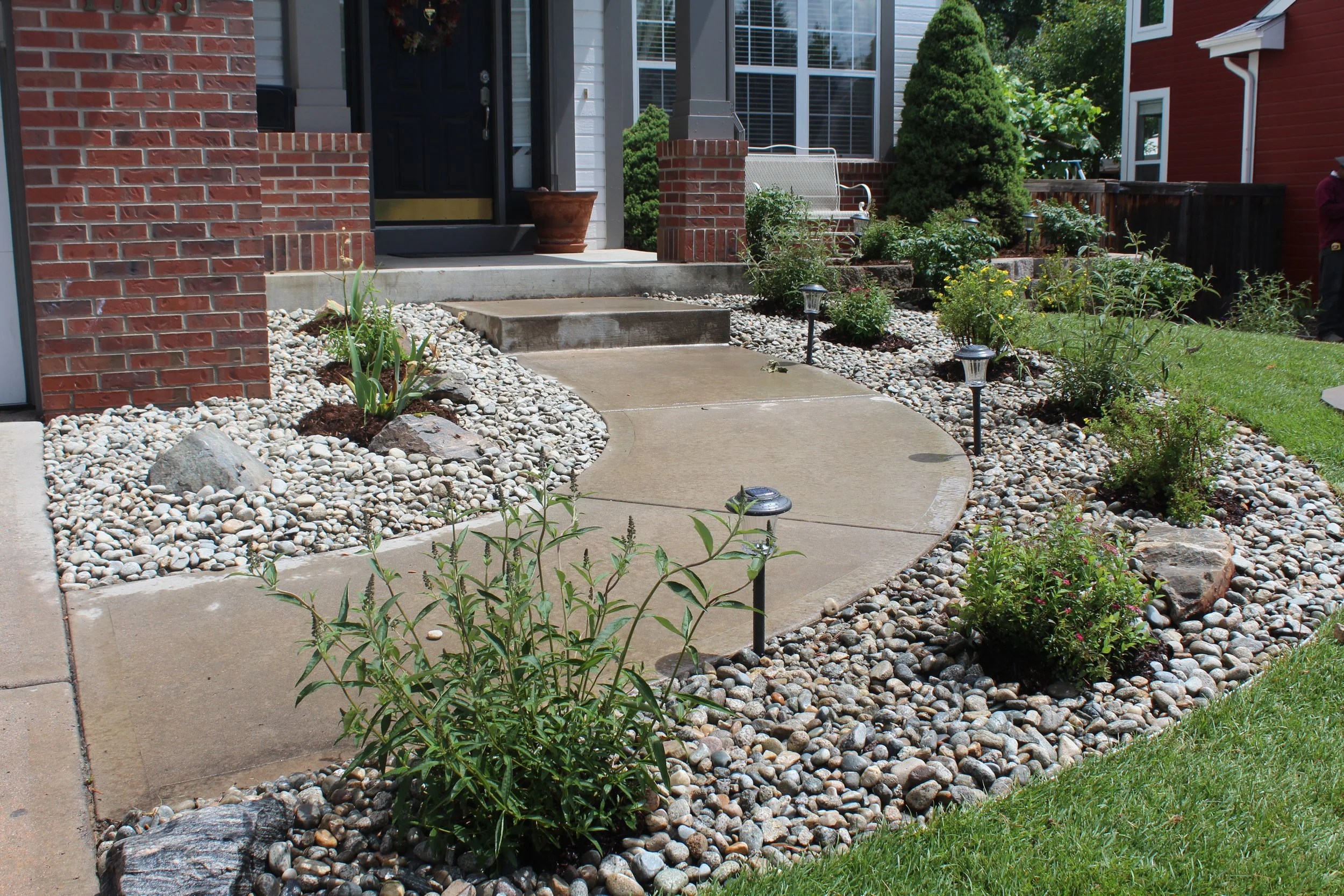 Front yard with a concrete walkway, small decorative rocks, and various green plants and shrubs, with a brick pillar and house entrance in the background.