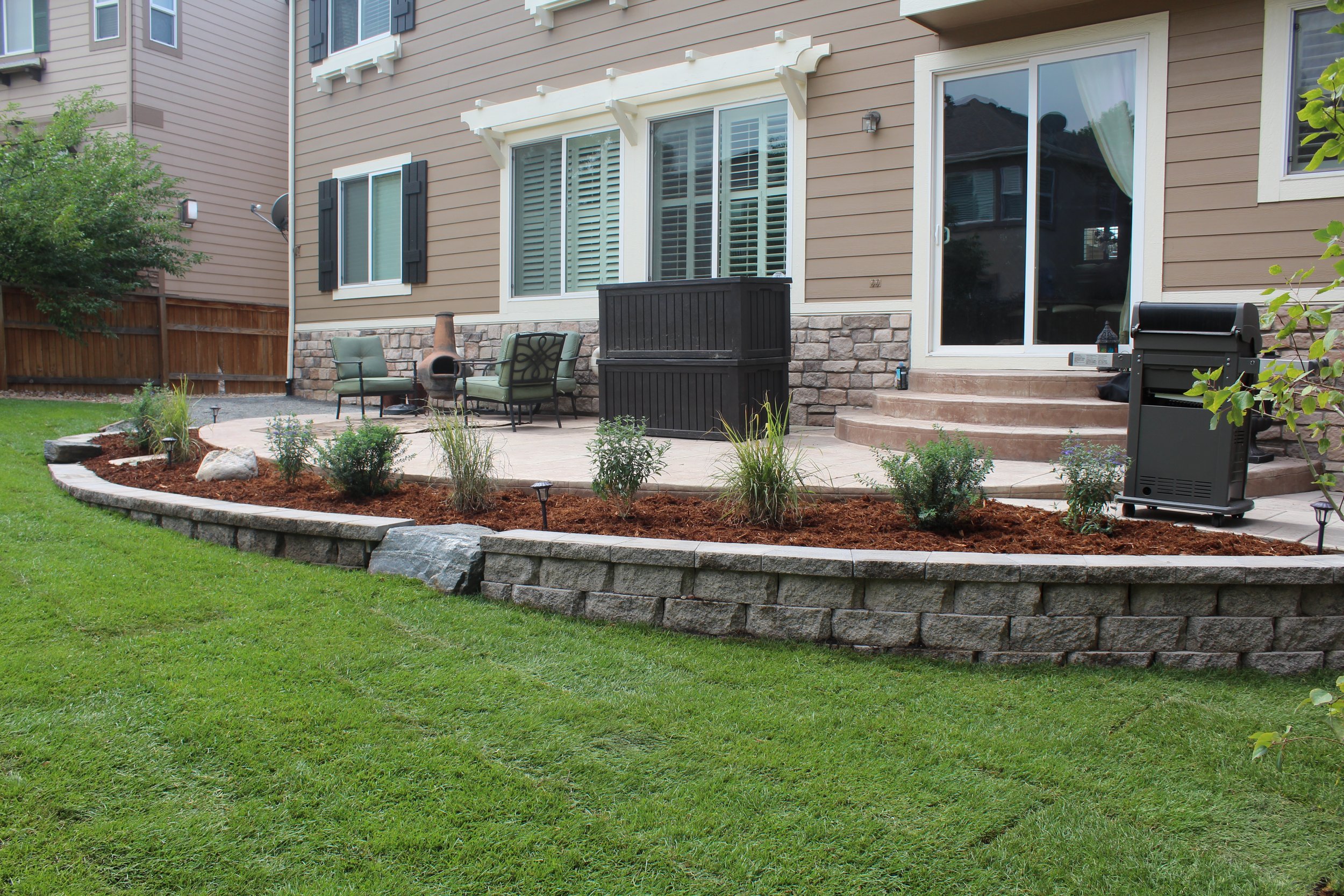 Retaining wall block and cap planter with small shrubs and mulch on the side of a concrete patio
