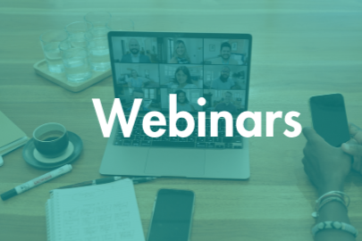 Laptop displaying a virtual meeting with multiple participants, with the word 'Webinars' overlaid on the screen. Items on the desk include a smartphone, open notebook, pen, cup of coffee, and a tray of glasses in the background.