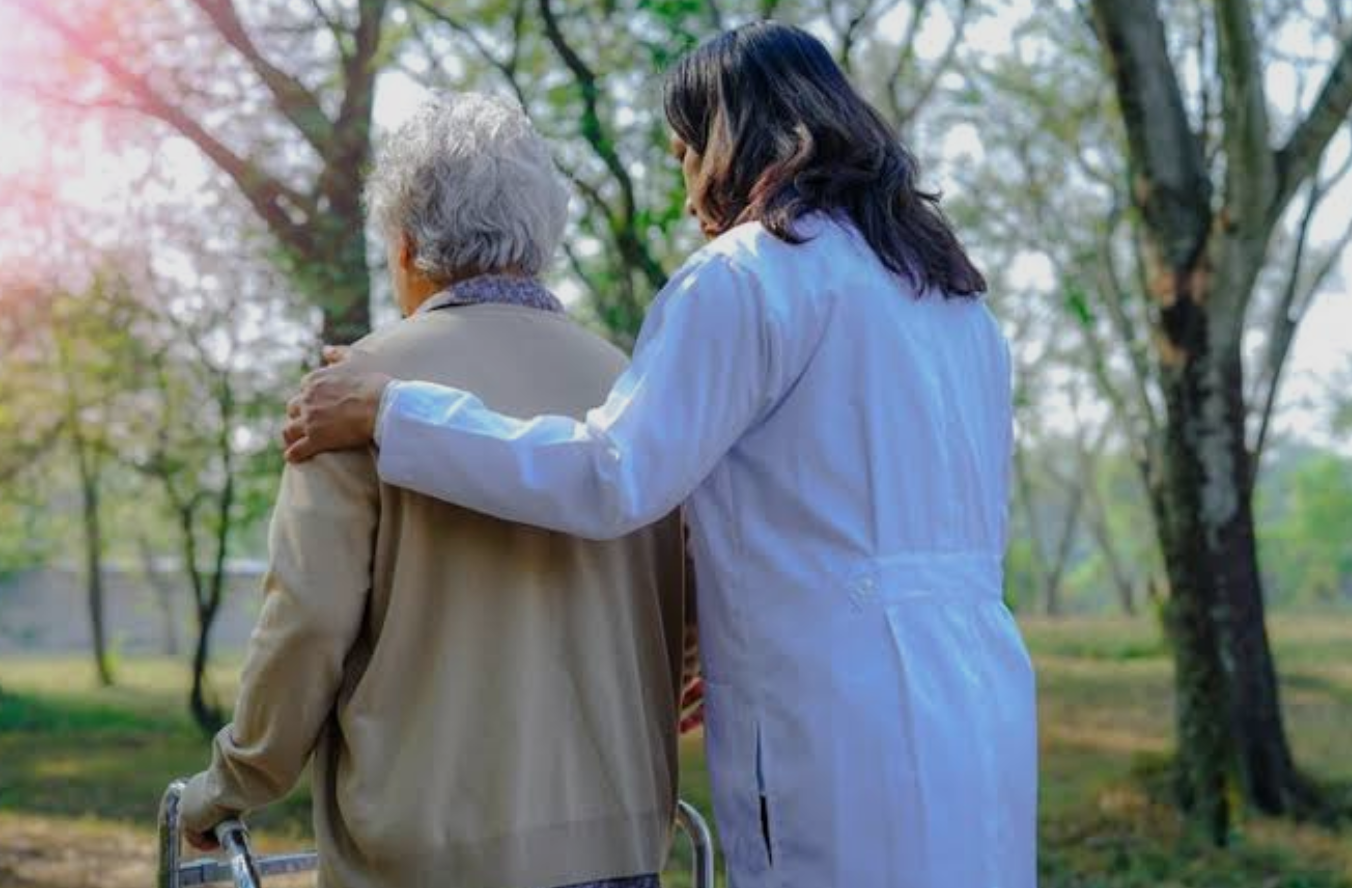 A woman in a white medical coat gently supports an elderly person with gray hair walking outdoors with a walker in a park surrounded by trees.