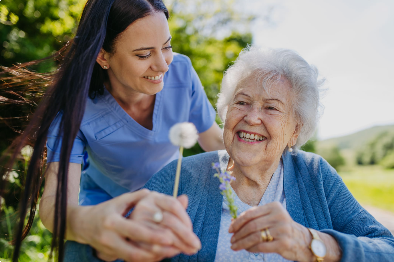 A young woman in light blue nursing scrubs smiling at an elderly woman with white hair, who is also smiling, outdoors on a sunny day with green trees in the background. The elderly woman is holding a small flower.