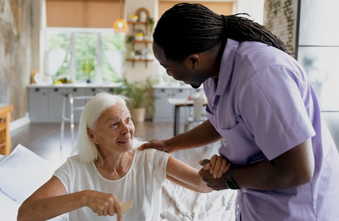 A caregiver helps an elderly woman in a nursing home or assisted living facility, holding her hand and gently supporting her.