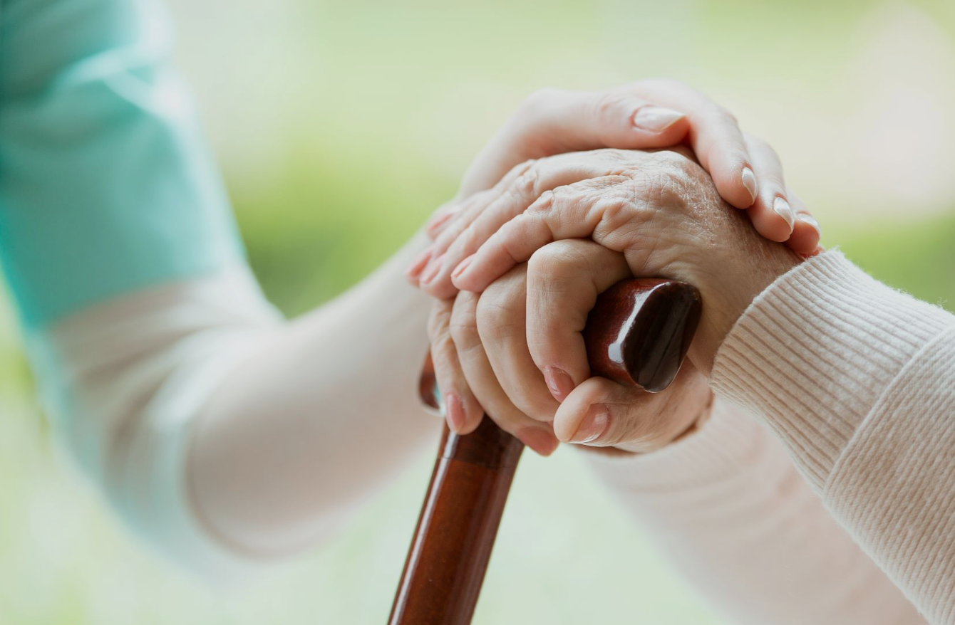 Two elderly people holding hands, one with a cane, with one hand on top of the other's and gently holding it