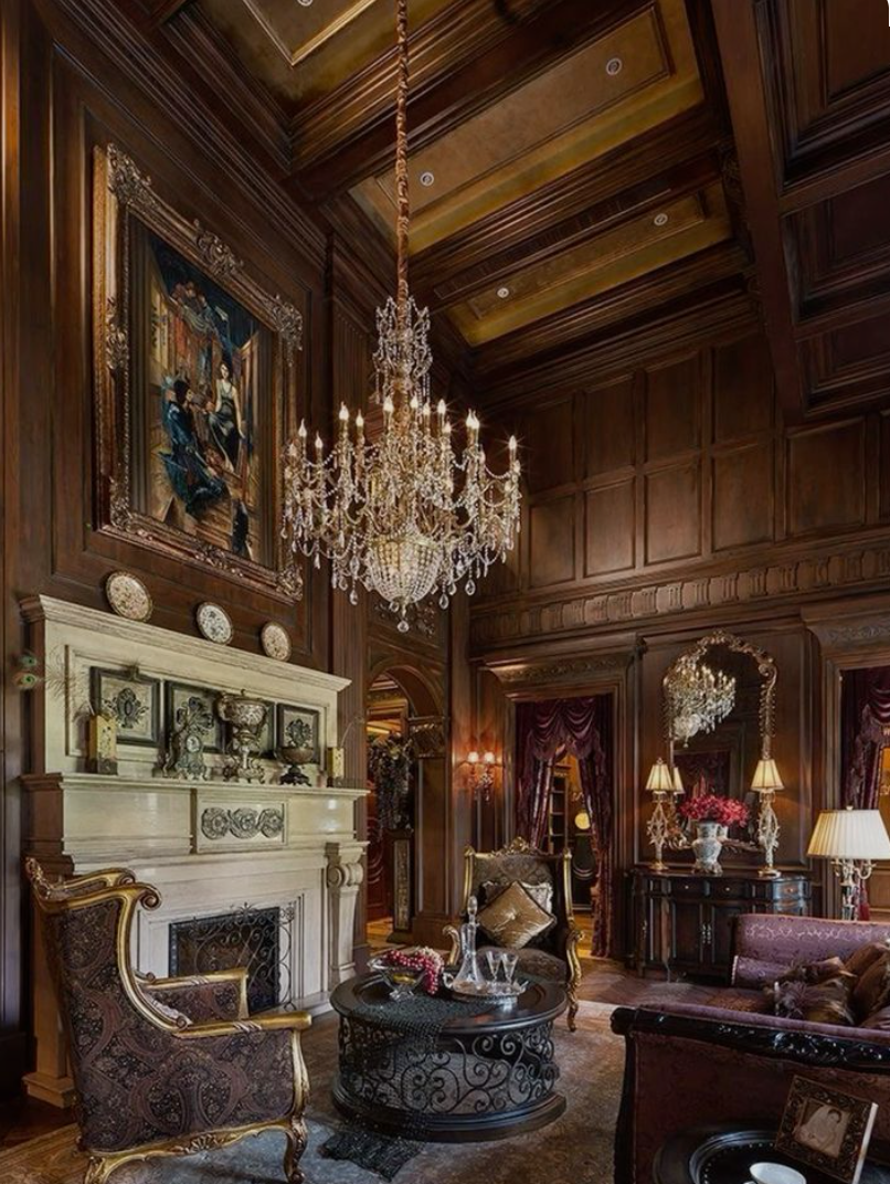 Luxurious living room with dark wood paneled walls, a large crystal chandelier, a fireplace with decorative plates on top, antique chairs, a round coffee table with drinks, and elegant lamps and curtains.