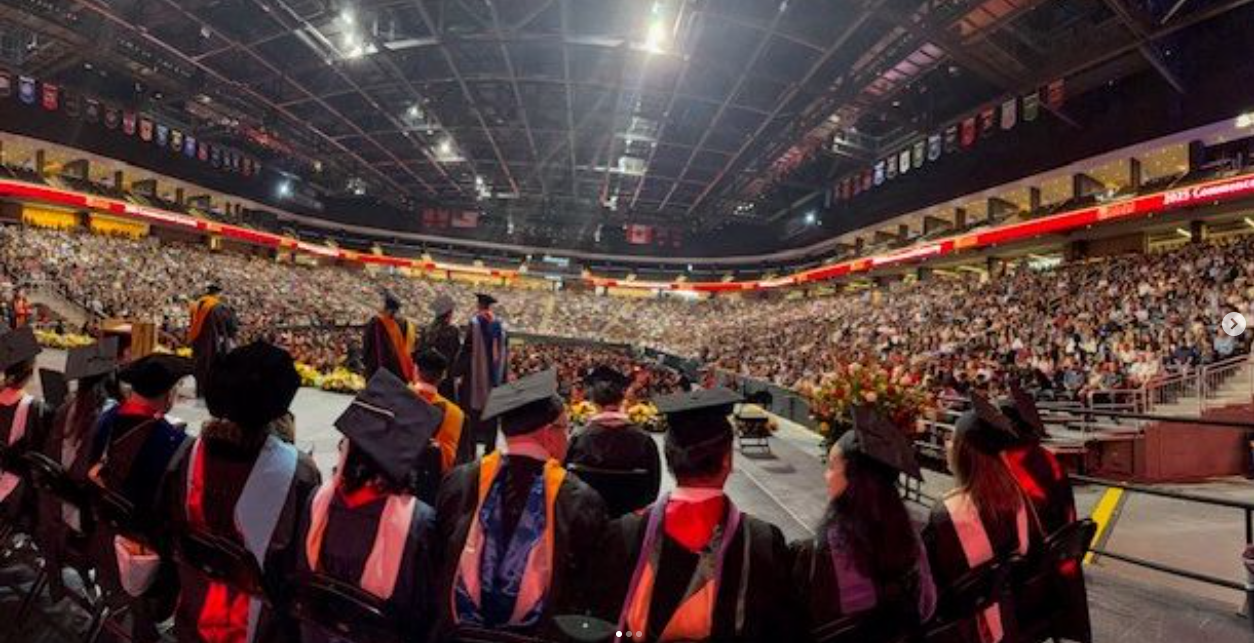 Graduates in caps and gowns seated in an indoor stadium with an audience in the stands, celebrating a graduation ceremony.