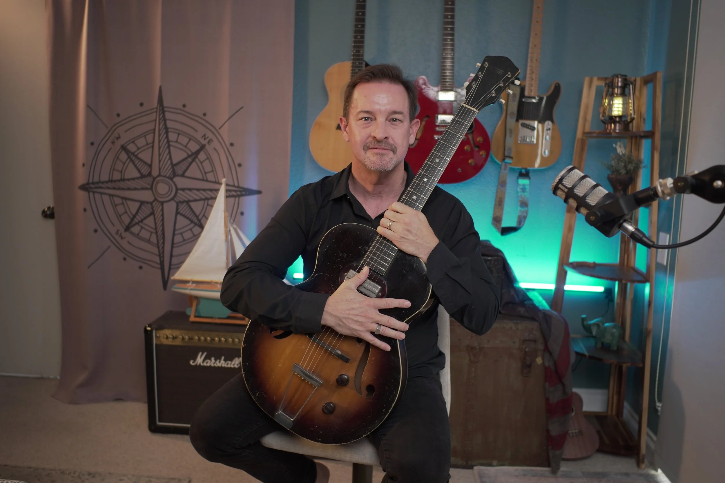 A man sitting on a stool in a music studio holding an acoustic guitar. Behind him are several other guitars hanging on the wall, a Marshall amplifier, a microphone, and various music-related decor."