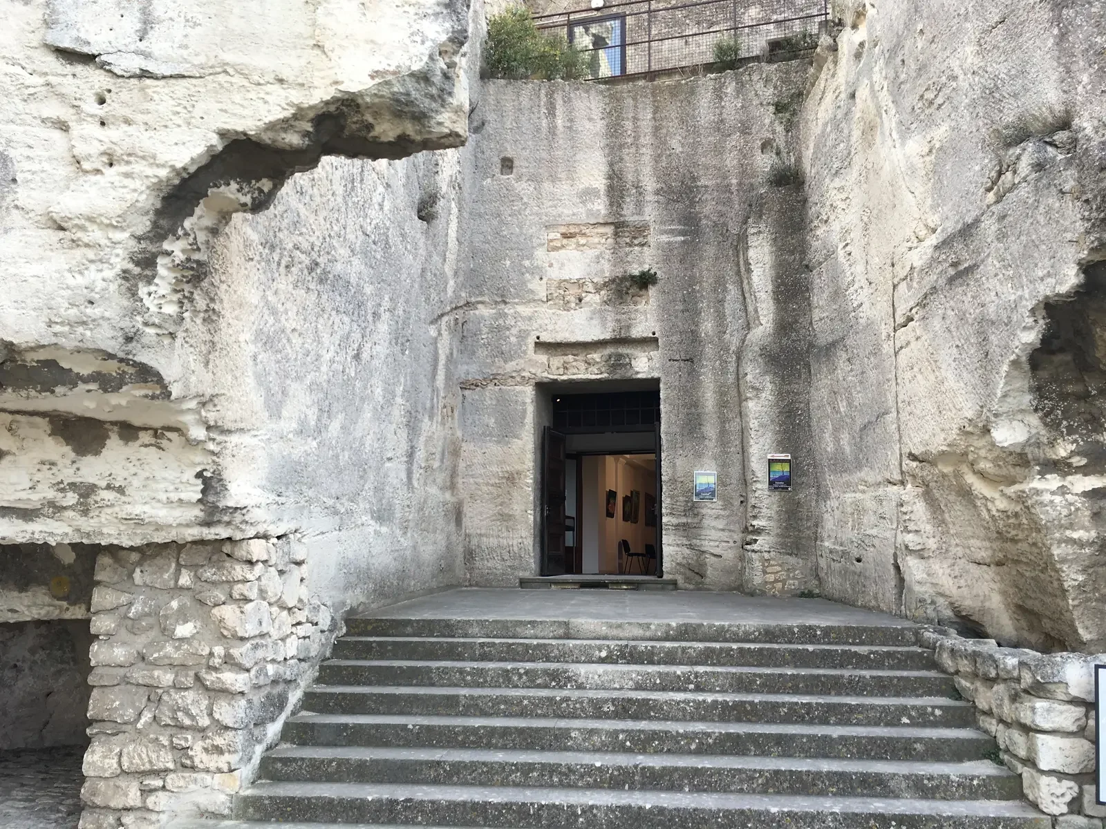 Entrée d'un musée ou d'une galerie d'art dans une structure rocheuse ancienne, avec des marches en pierre menant à la porte ouverte, à l'intérieur on voit des œuvres d'art sur les murs.