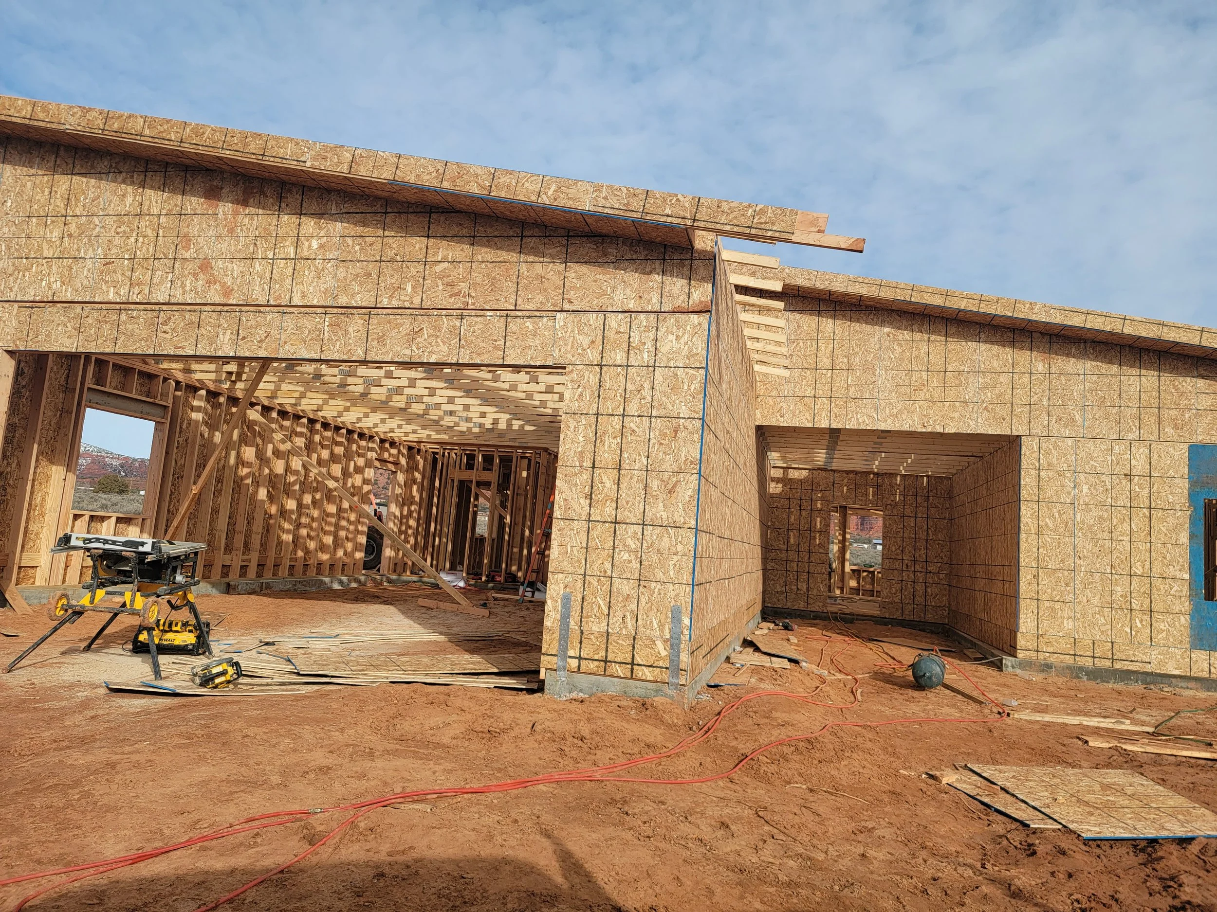 Construction site with wooden framing and plywood walls, power tools, construction materials, and electrical cords on the ground, under a blue sky.