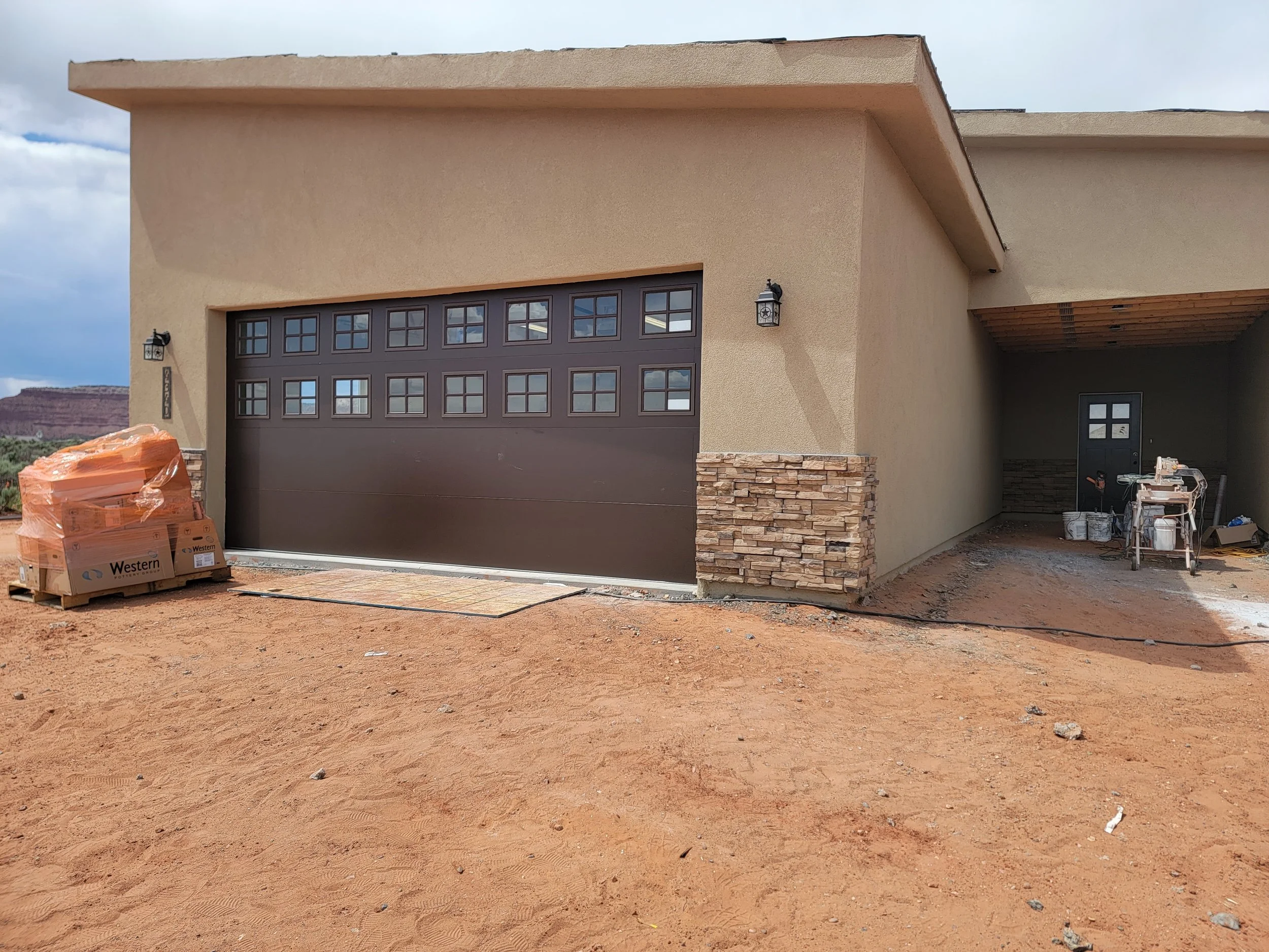 Front view of a house under construction with a closed garage door, beige stucco walls, stone accents at the base, and construction materials nearby, with a cloudy sky in the background.