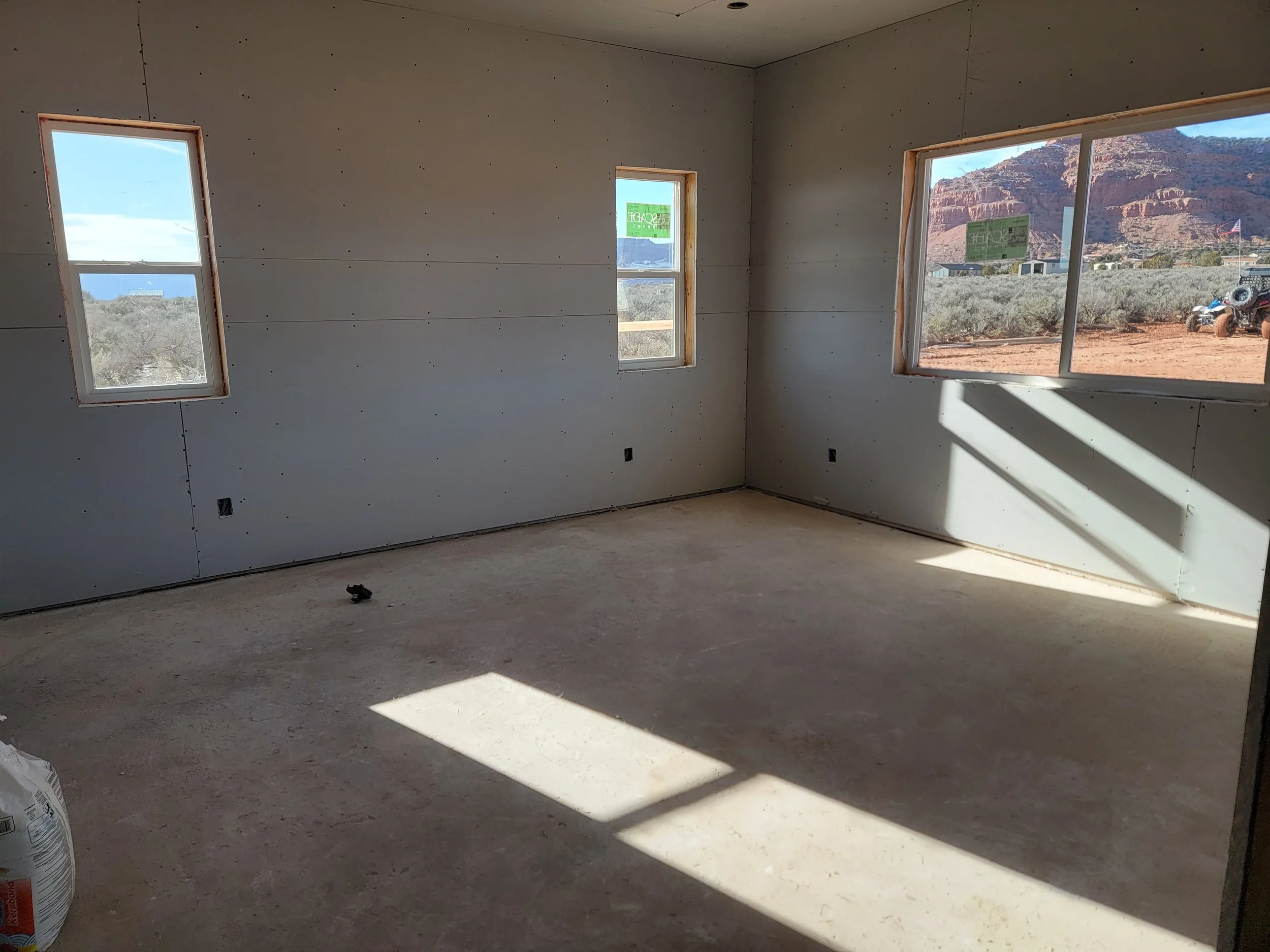 Interior of a building under construction with unfinished walls and four windows, showing an outdoor desert landscape with red rocks, truck, and American flag.