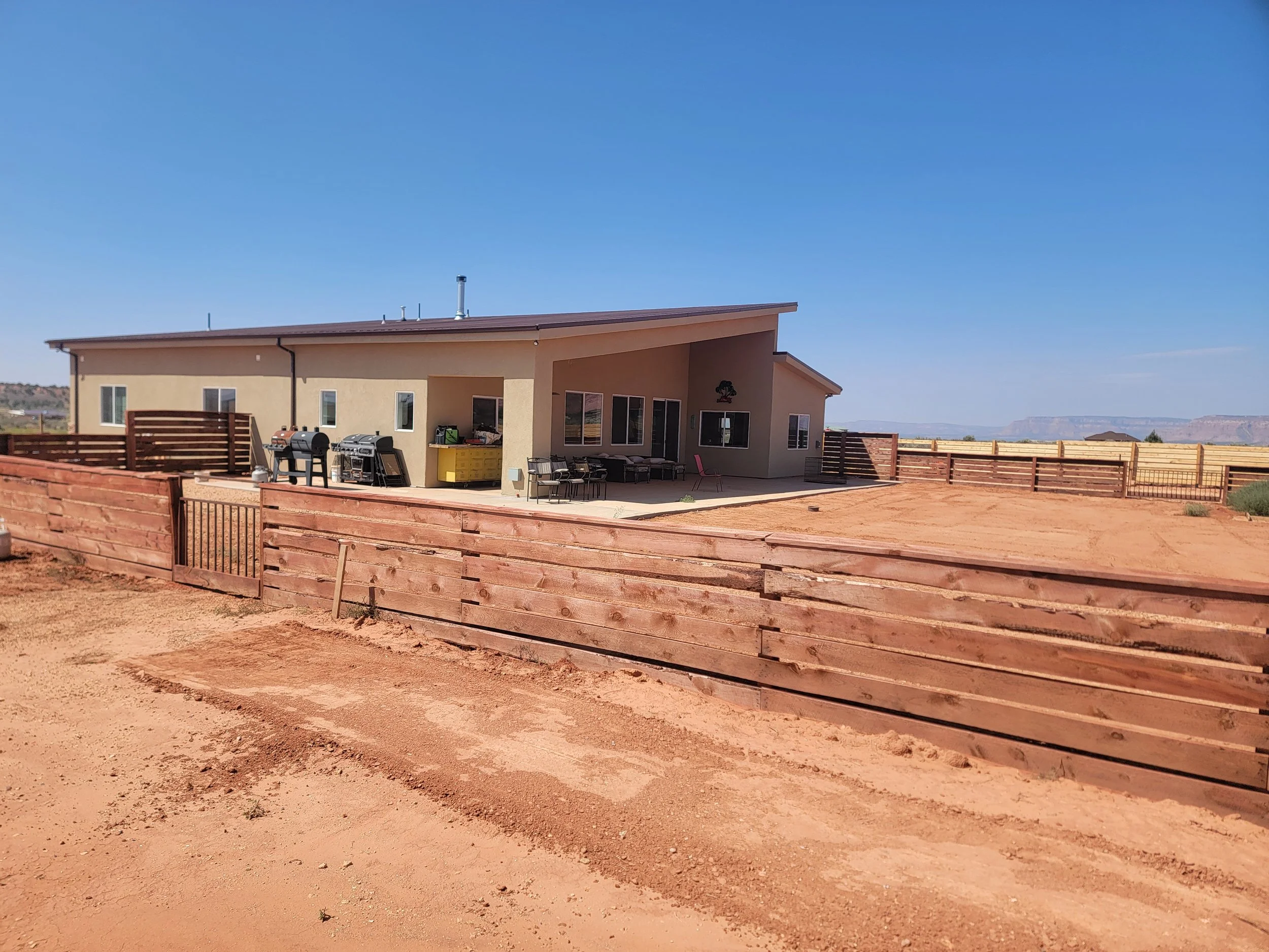 A house with a patio area, surrounded by a wooden fence, in a landscape with distant mesas and a clear blue sky.
