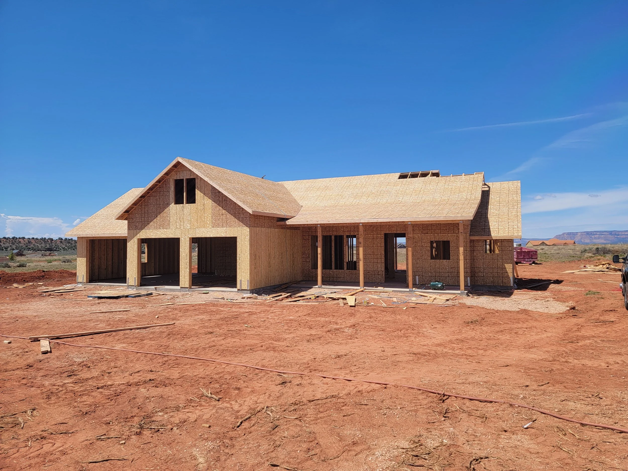 A house under construction with wooden framing and roof in an open landscape under a clear blue sky.