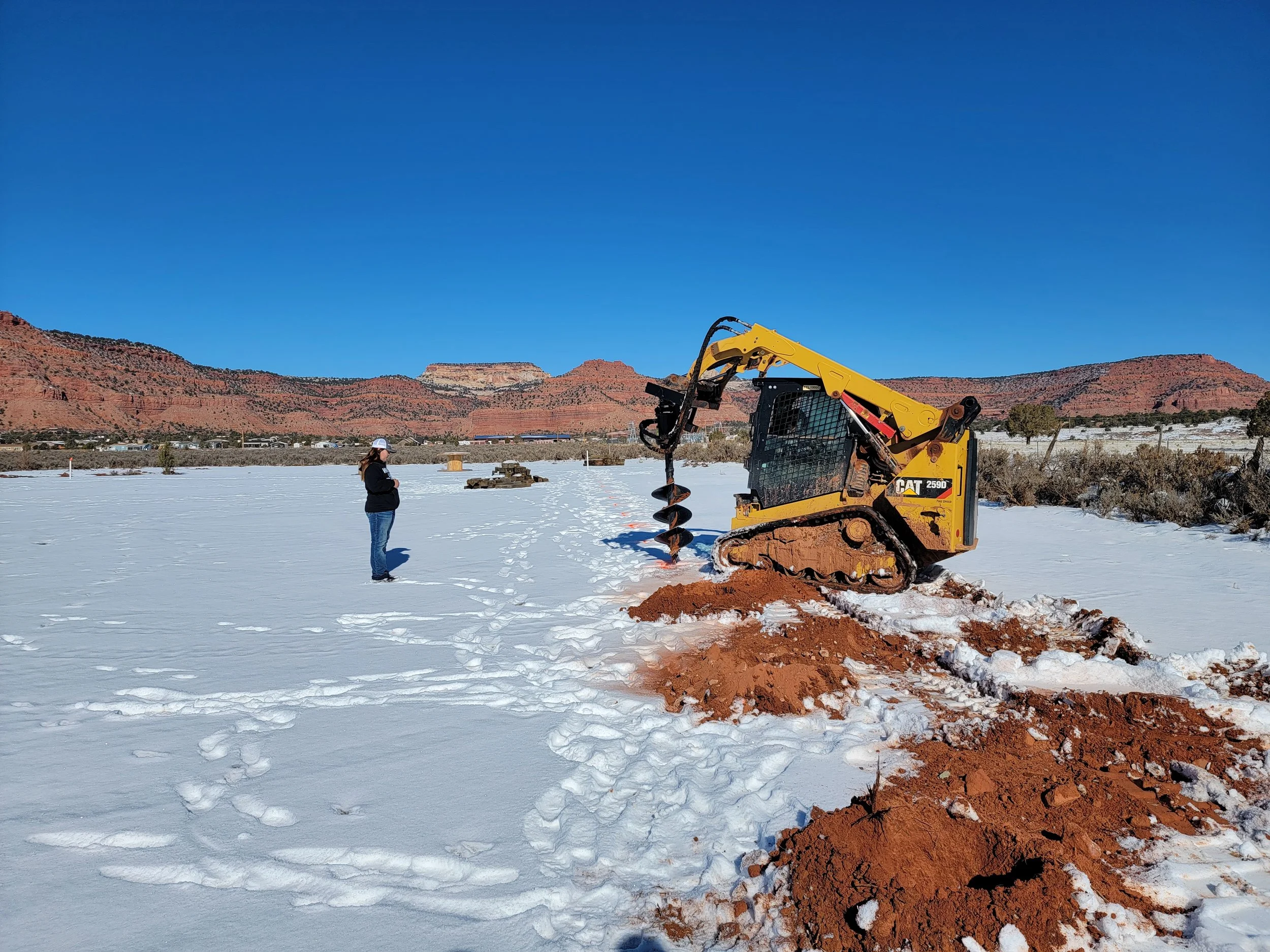 A person standing on snow-covered ground near a yellow Caterpillar excavator with a large auger attachment, with red rock formations and mountains visible in the background under a clear blue sky.