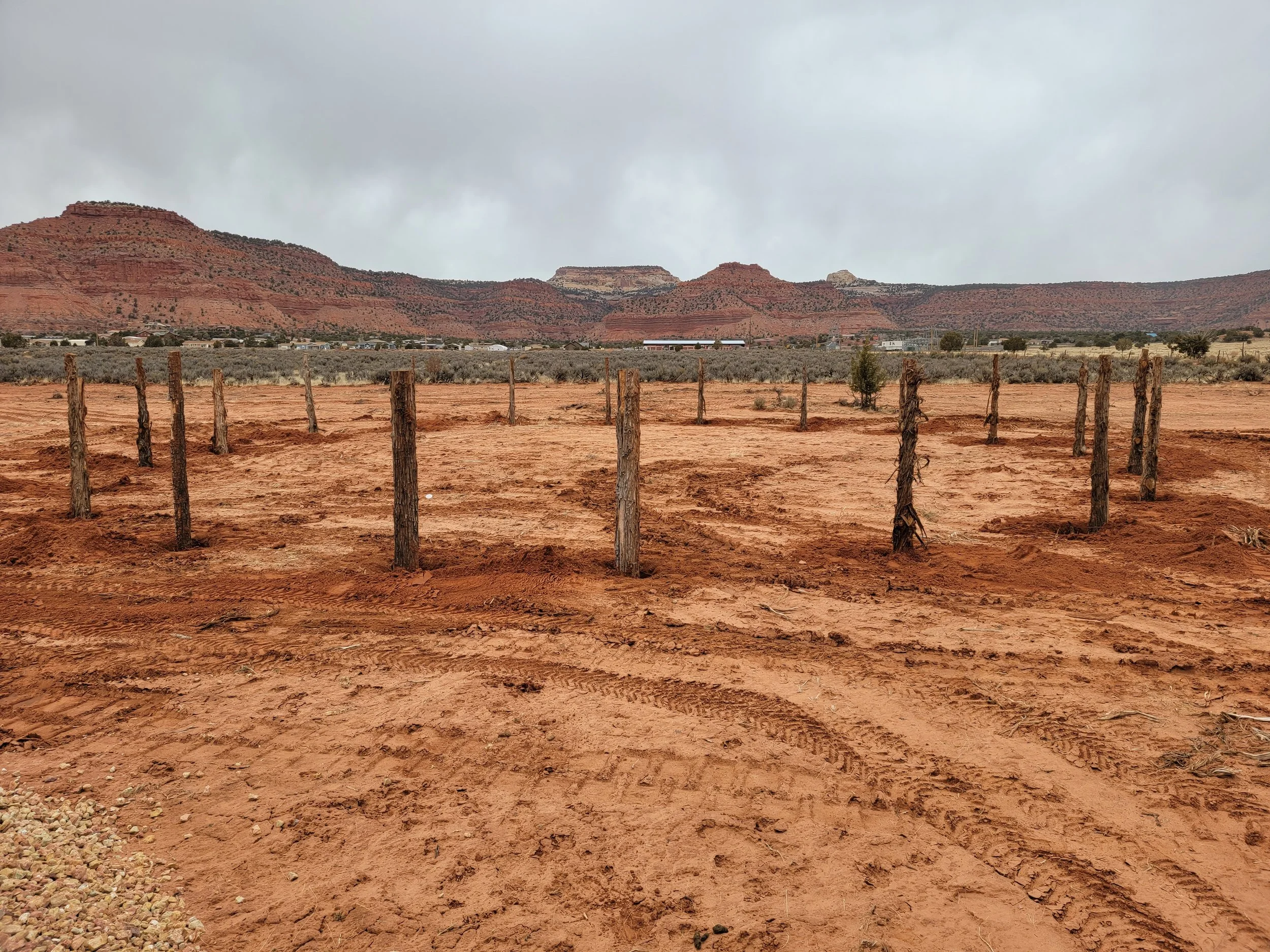 Desert landscape with reddish-brown rock formations and cloudy sky, sparse vegetation, and wooden fence posts in the foreground.