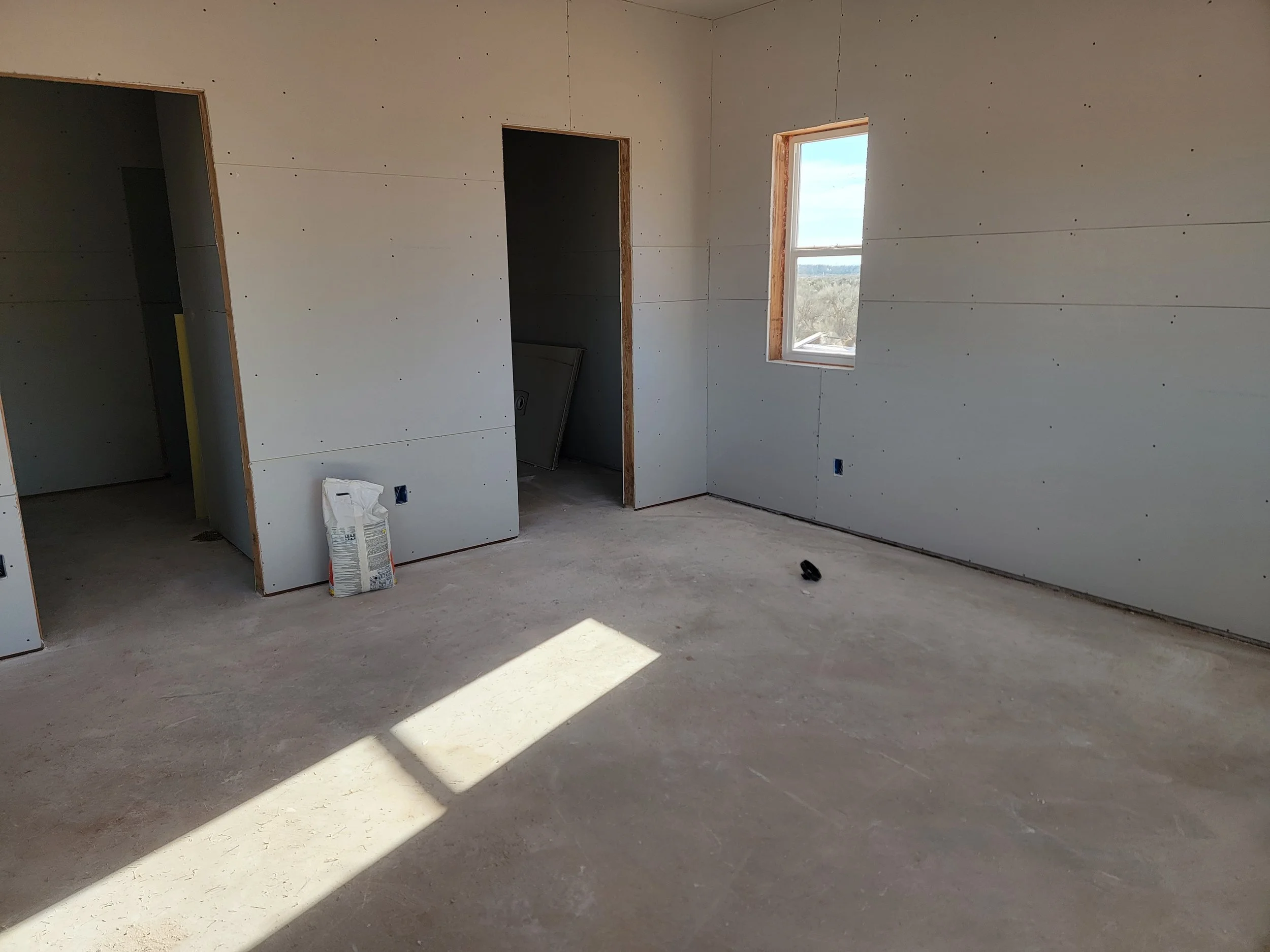 An unfinished room with sheetrock walls, a window letting in sunlight, and visible construction materials on the floor.