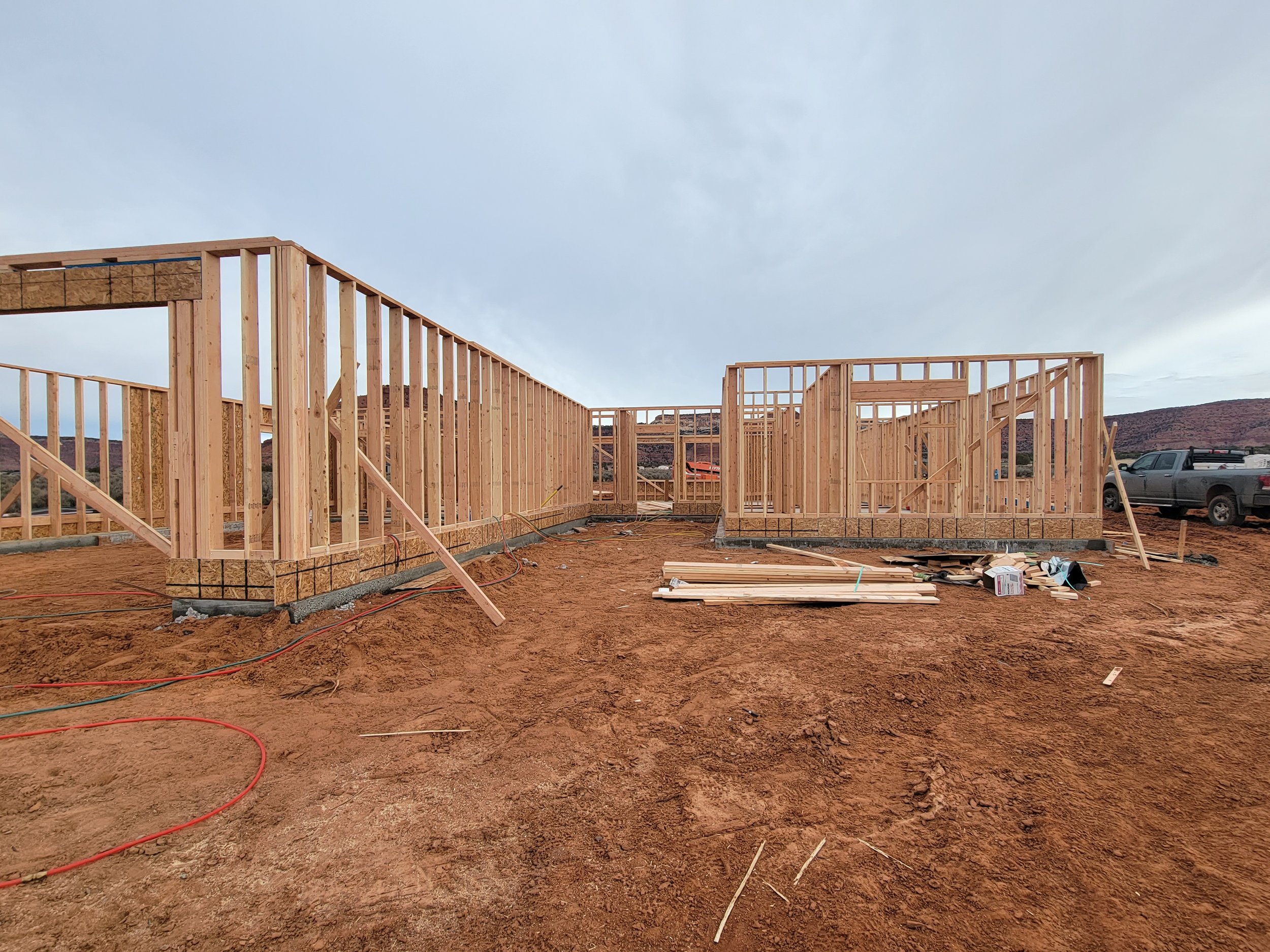Construction site with wooden framing for a building, construction materials, and vehicles in an open area with a mountainous background under a cloudy sky.