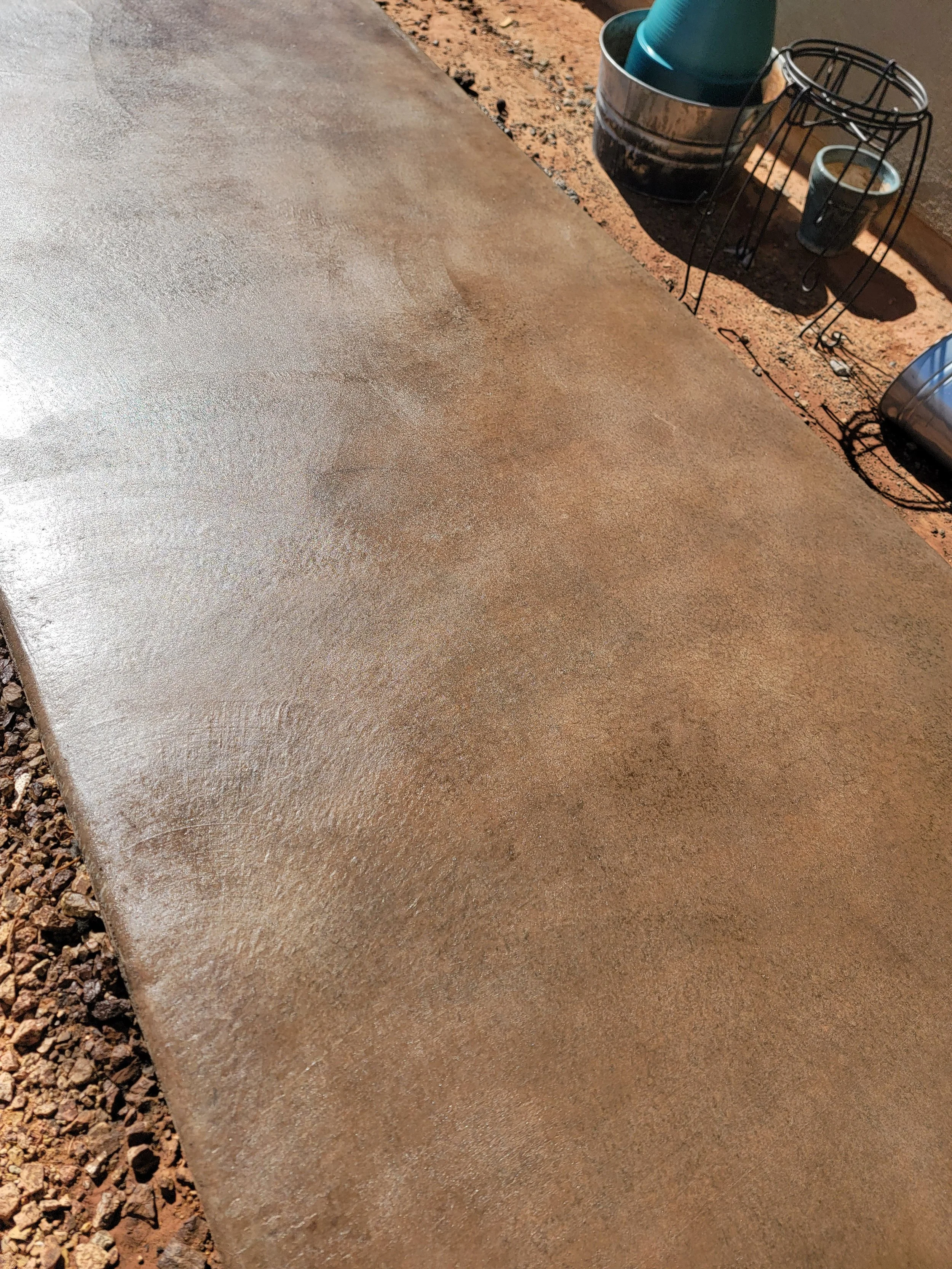 Close-up of a freshly poured concrete sidewalk with a slightly textured surface, adjacent to a gravel area on the left, and a section of metal tool or container with buckets and cups in the background.