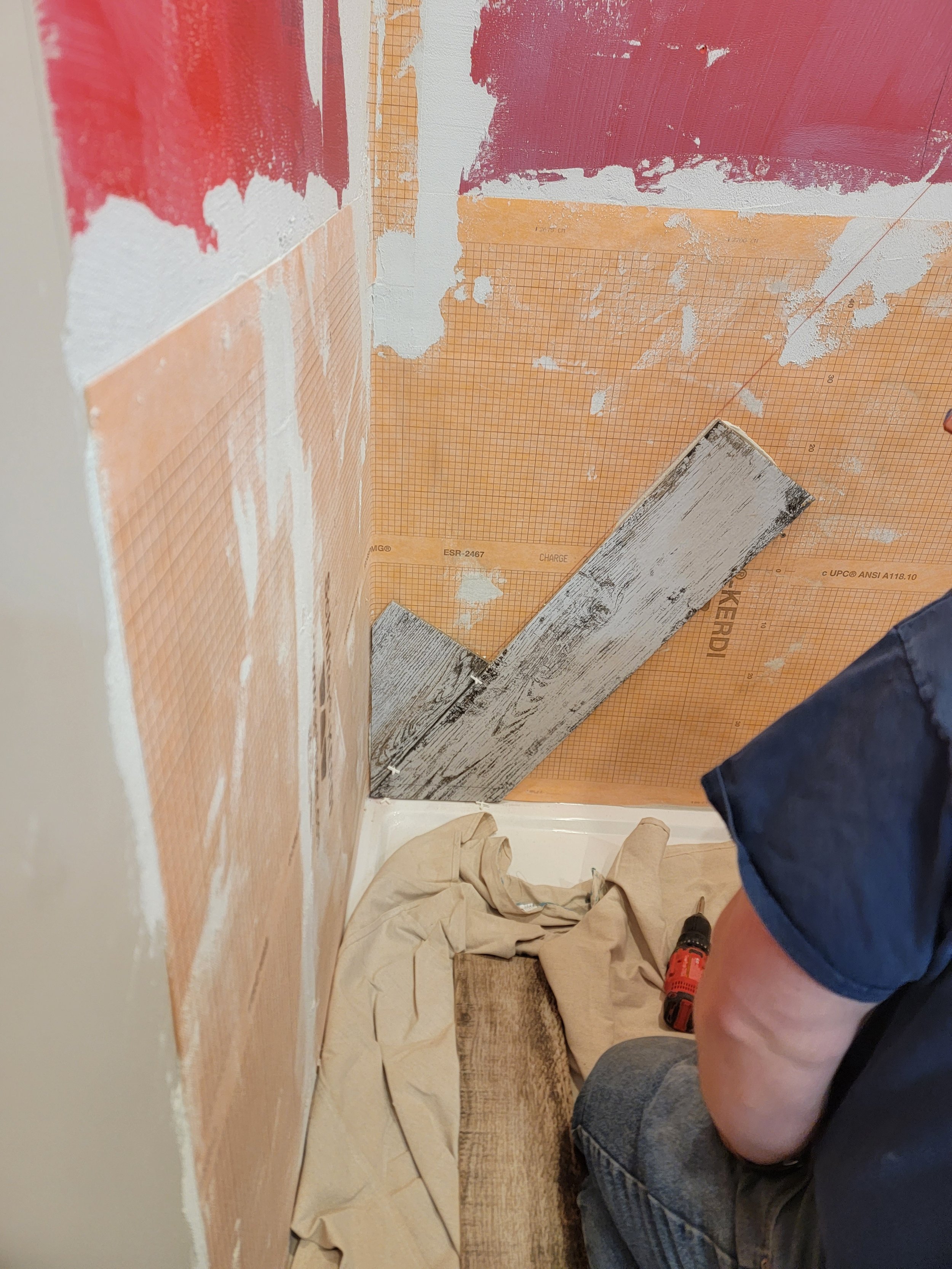 Person working on wall painting or renovation, with a drill in hand, wearing shorts and a sleeveless shirt, and a tarp on the floor.