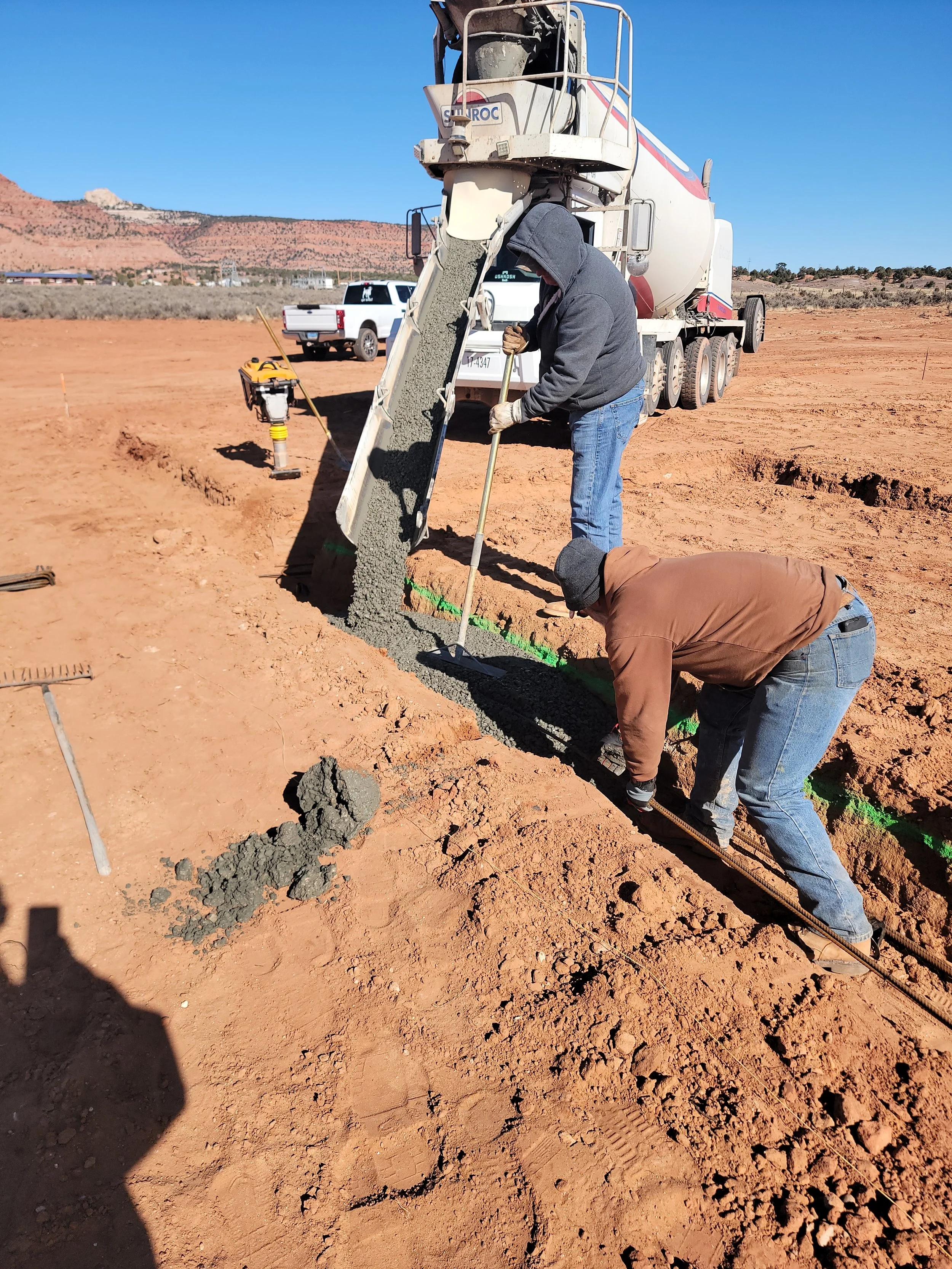 Two construction workers, one standing and one crouching, are pouring and leveling wet concrete into a trench at a construction site with a cement mixer truck nearby.