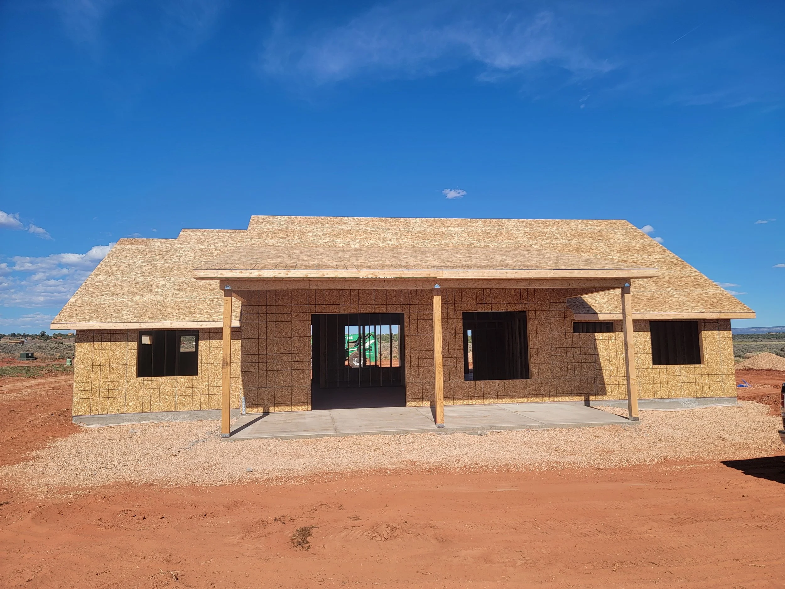 Front view of a house under construction with a sloped roof and open windows, set against a bright blue sky with scattered clouds.