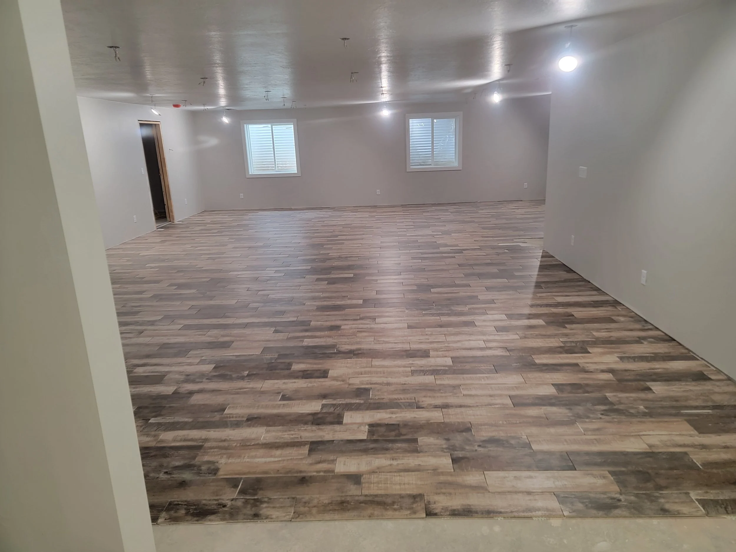 Empty room with white walls, ceiling lights, two windows, and wood-like flooring, with an open doorway on the left side.