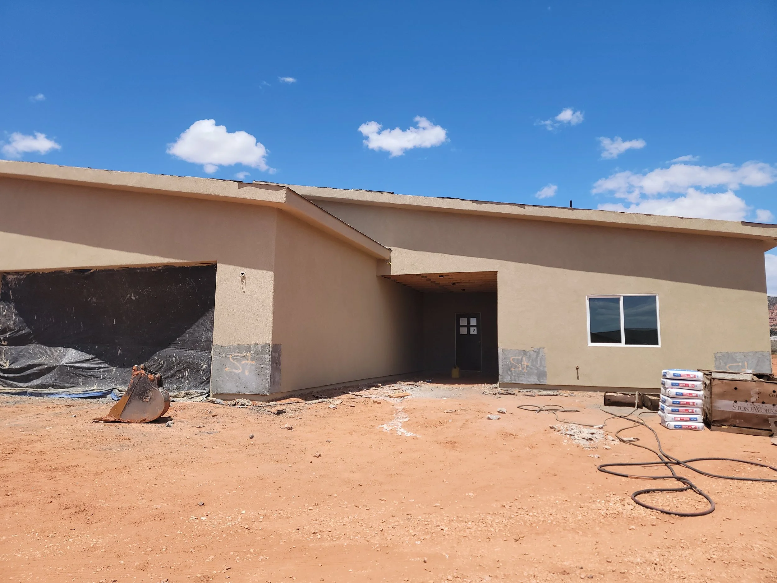 Under construction house with beige exterior walls and large windows, construction materials and tools on bare dirt ground, blue sky with scattered clouds overhead.