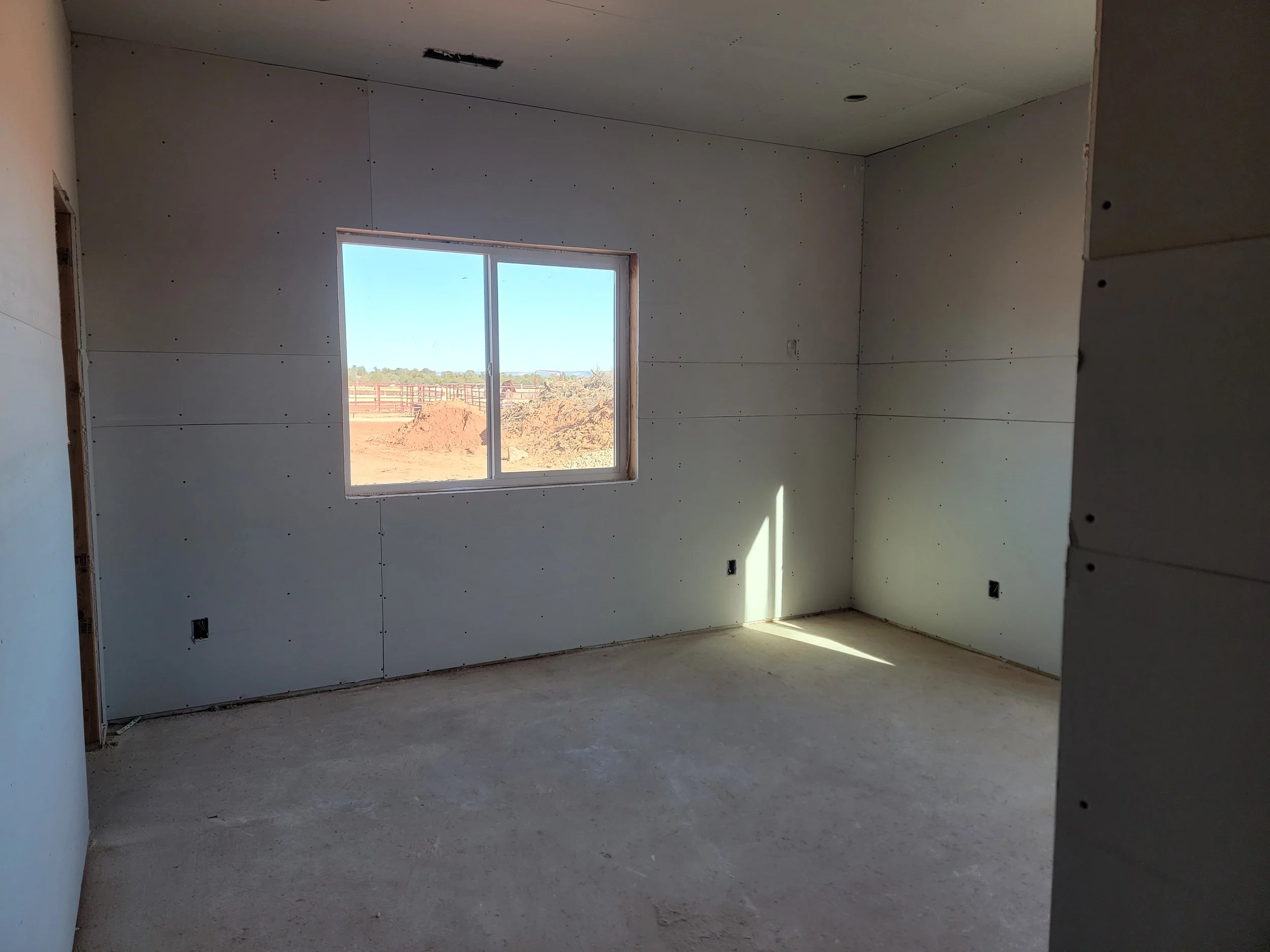 Interior of a room under construction with drywall installed and a window showing an outdoor view of a yard and distant trees.