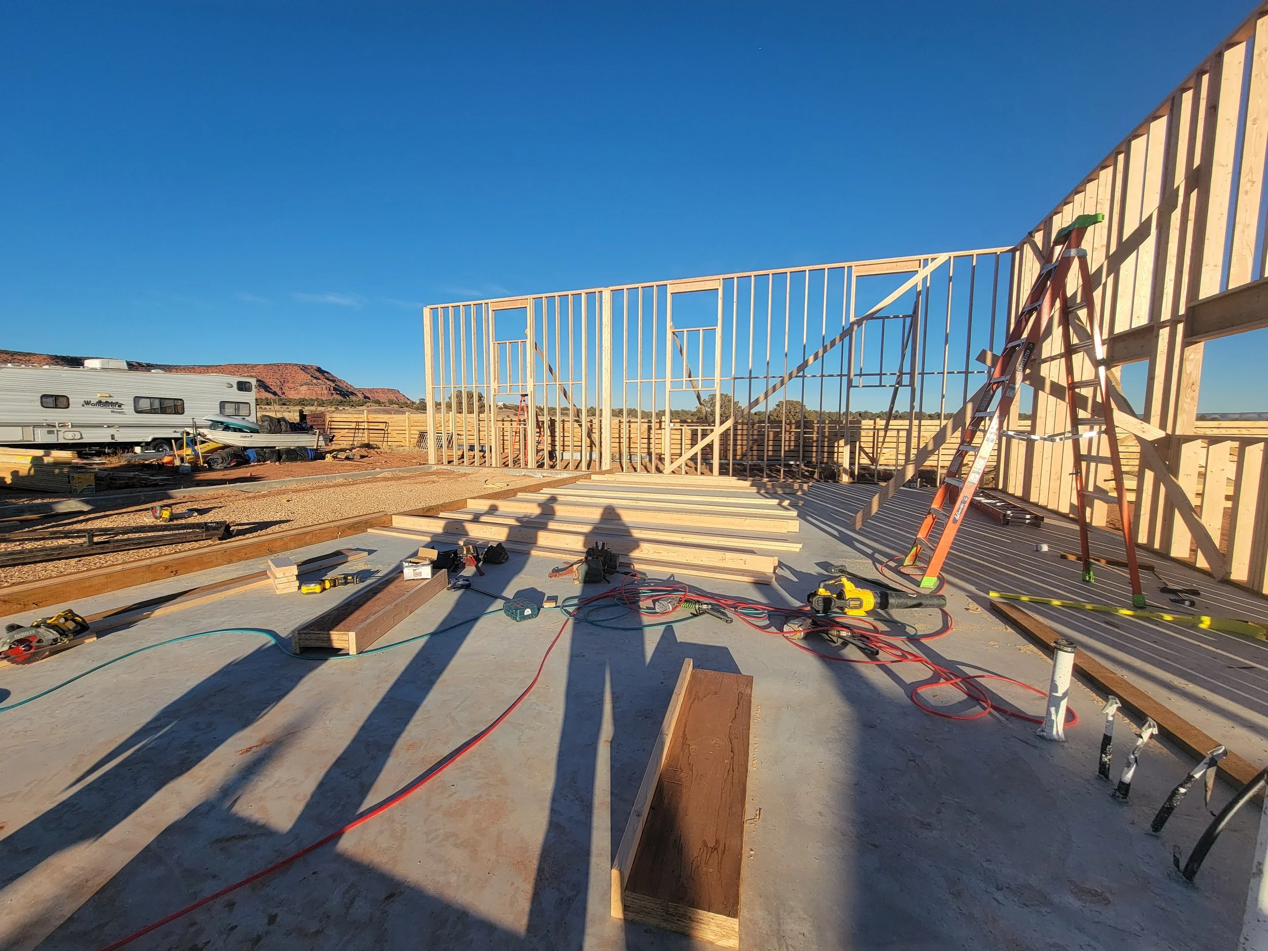 Construction site with wooden framing for a building, various tools and materials scattered on the ground, and a ladder leaning against the structure. In the background, there are RVs, a fence, and distant rock formations under a clear blue sky.