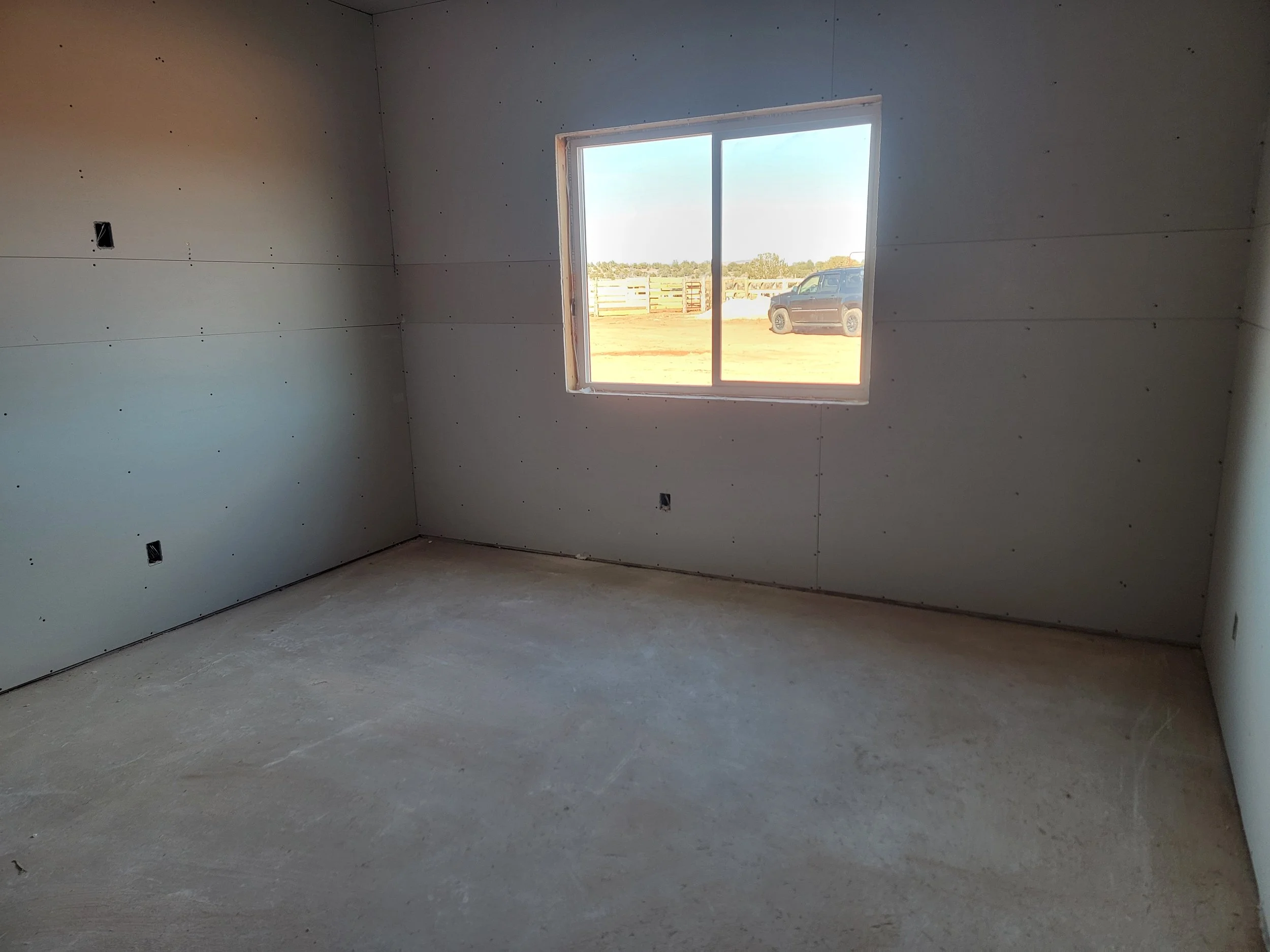 Interior of a room under construction with unpainted drywall walls and floor, a window showing an outdoor scene with a vehicle and wooden fence.