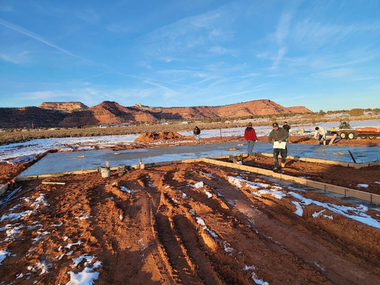 Construction workers laying concrete foundation on a muddy site with mountains in the background under a blue sky.