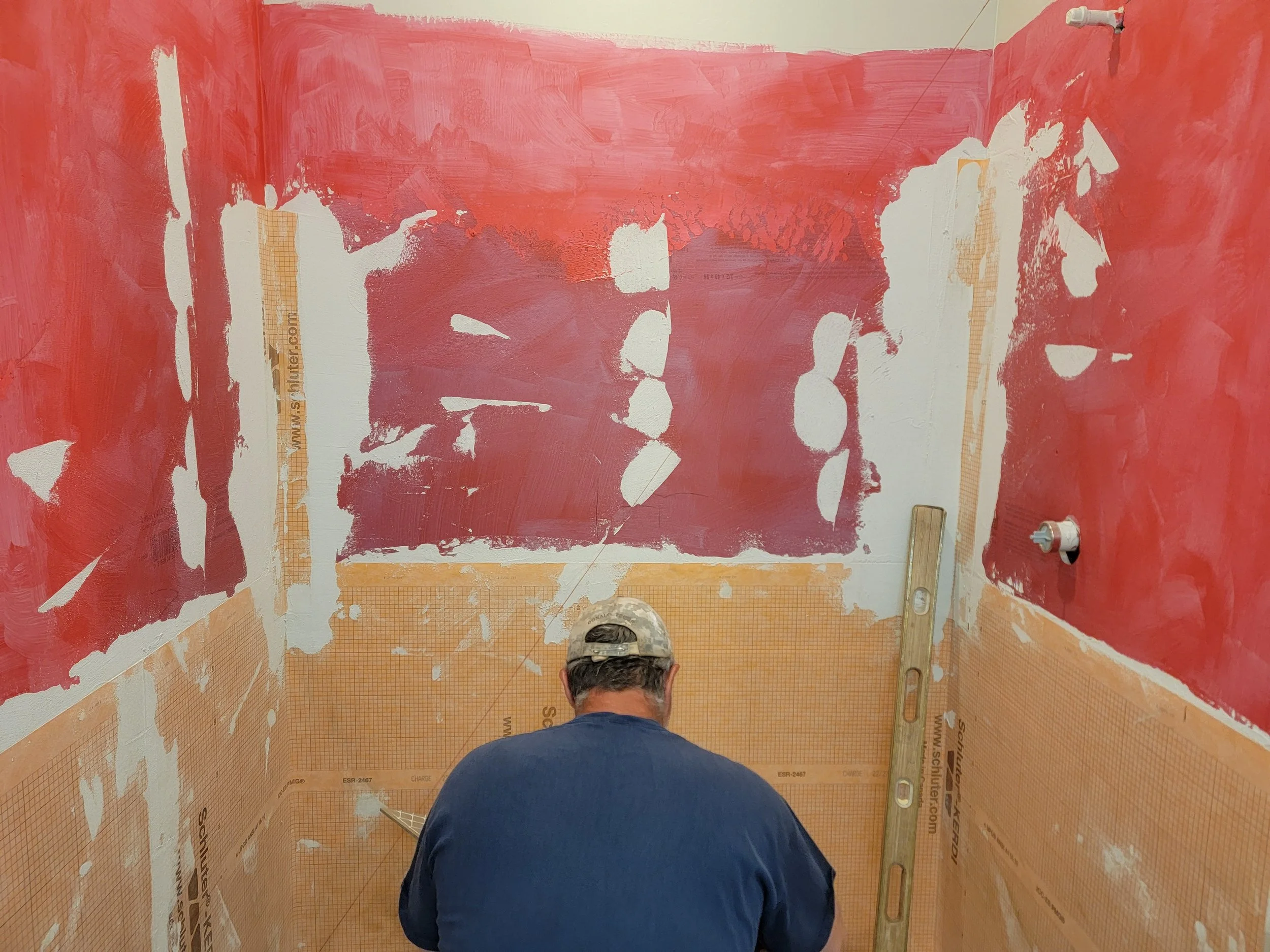 A person in a baseball cap and blue shirt standing in a small room with red paint on the walls, which are partly covered with orange protective material, preparing to paint or drywall.