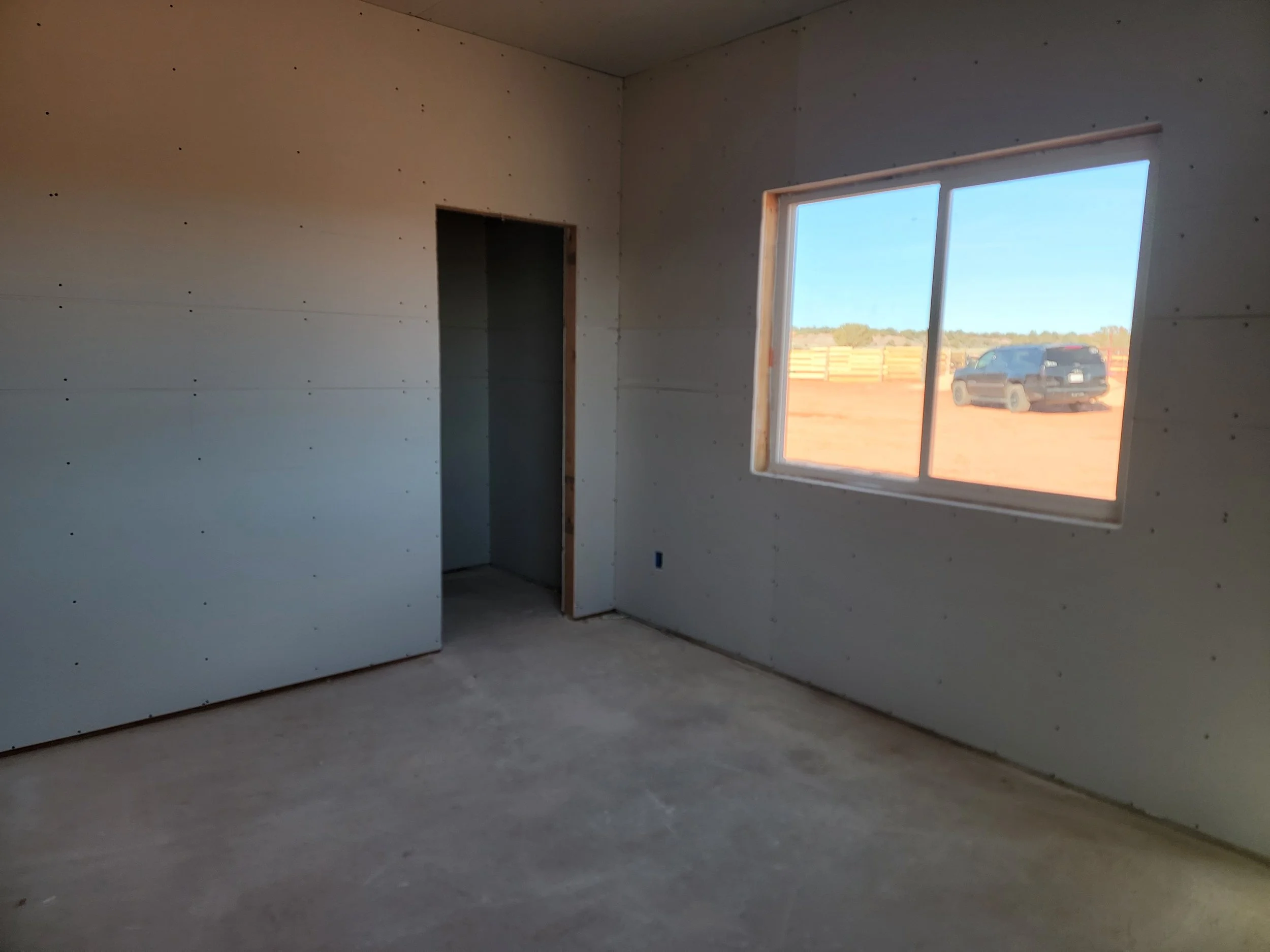 Empty room under construction with unfinished drywall walls, a window allowing natural light, and a view of a parked vehicle outside.