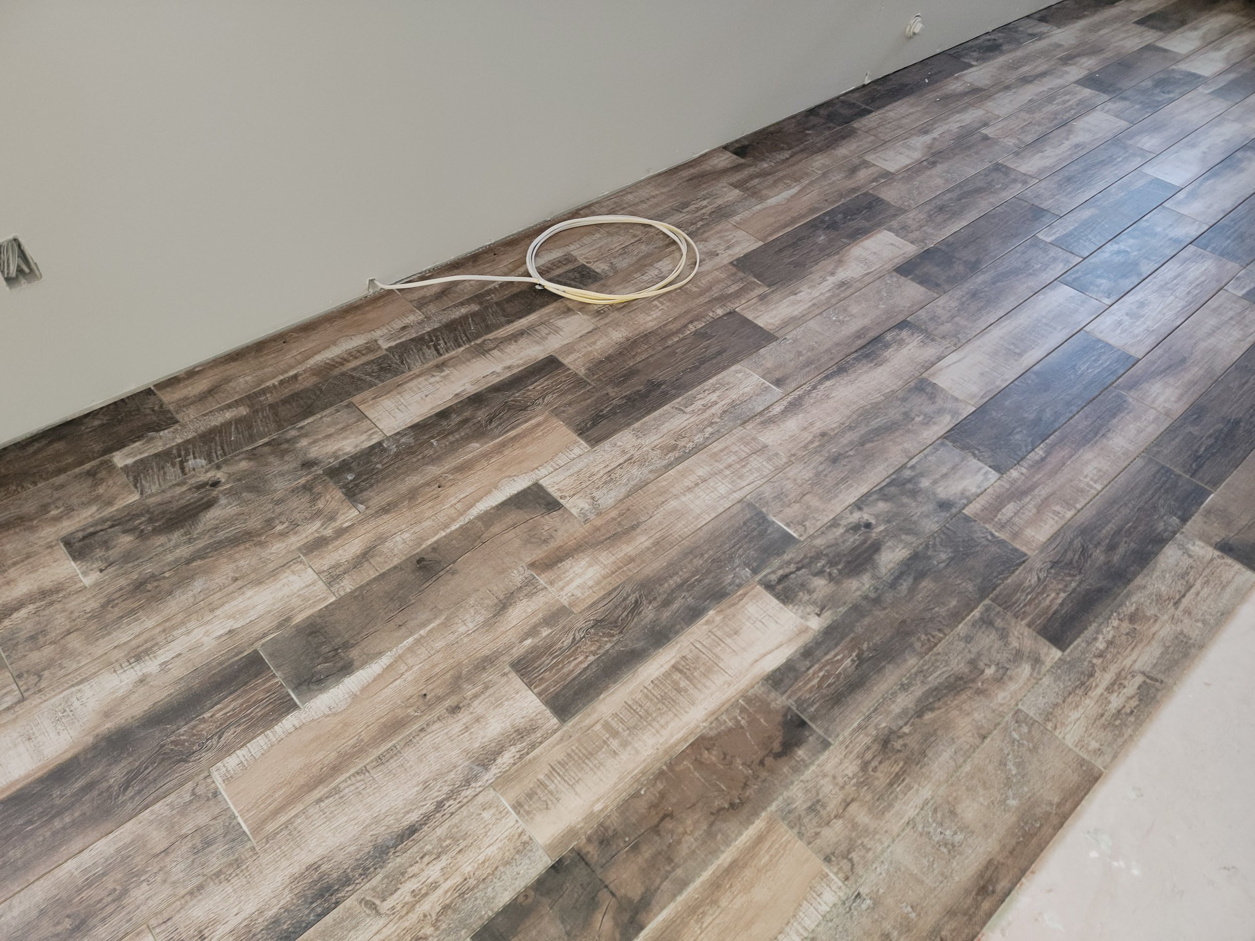 A section of a room with a brown wood-look tile floor, an unfinished light gray wall, and a loose white cable on the floor.