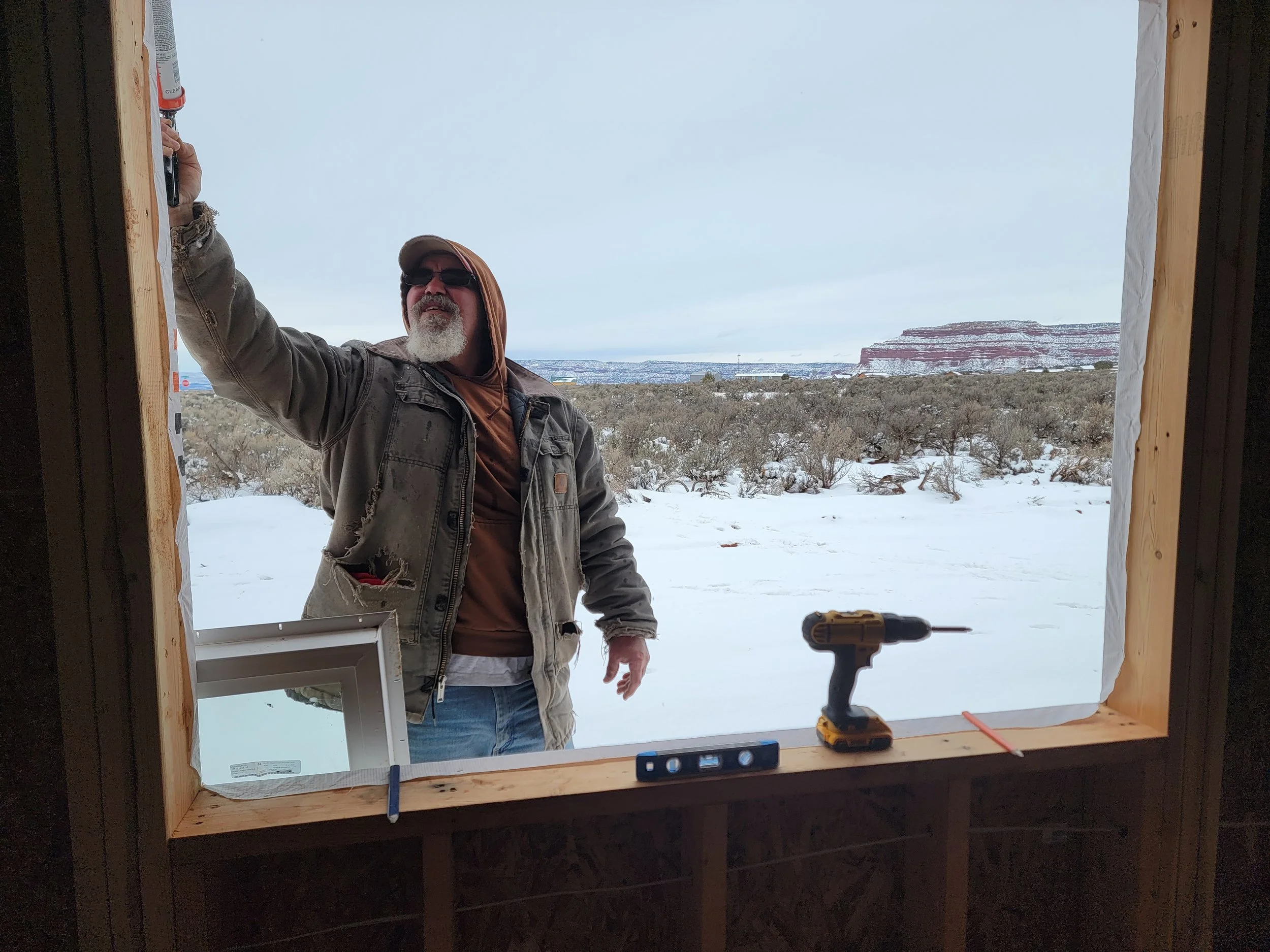 A man with a white beard, sunglasses, a brown hoodie, and a beige jacket is standing outdoors in a snowy landscape, holding a clipboard. The view through the window frame shows snow-covered ground, leafless bushes, and a layered red rock formation in