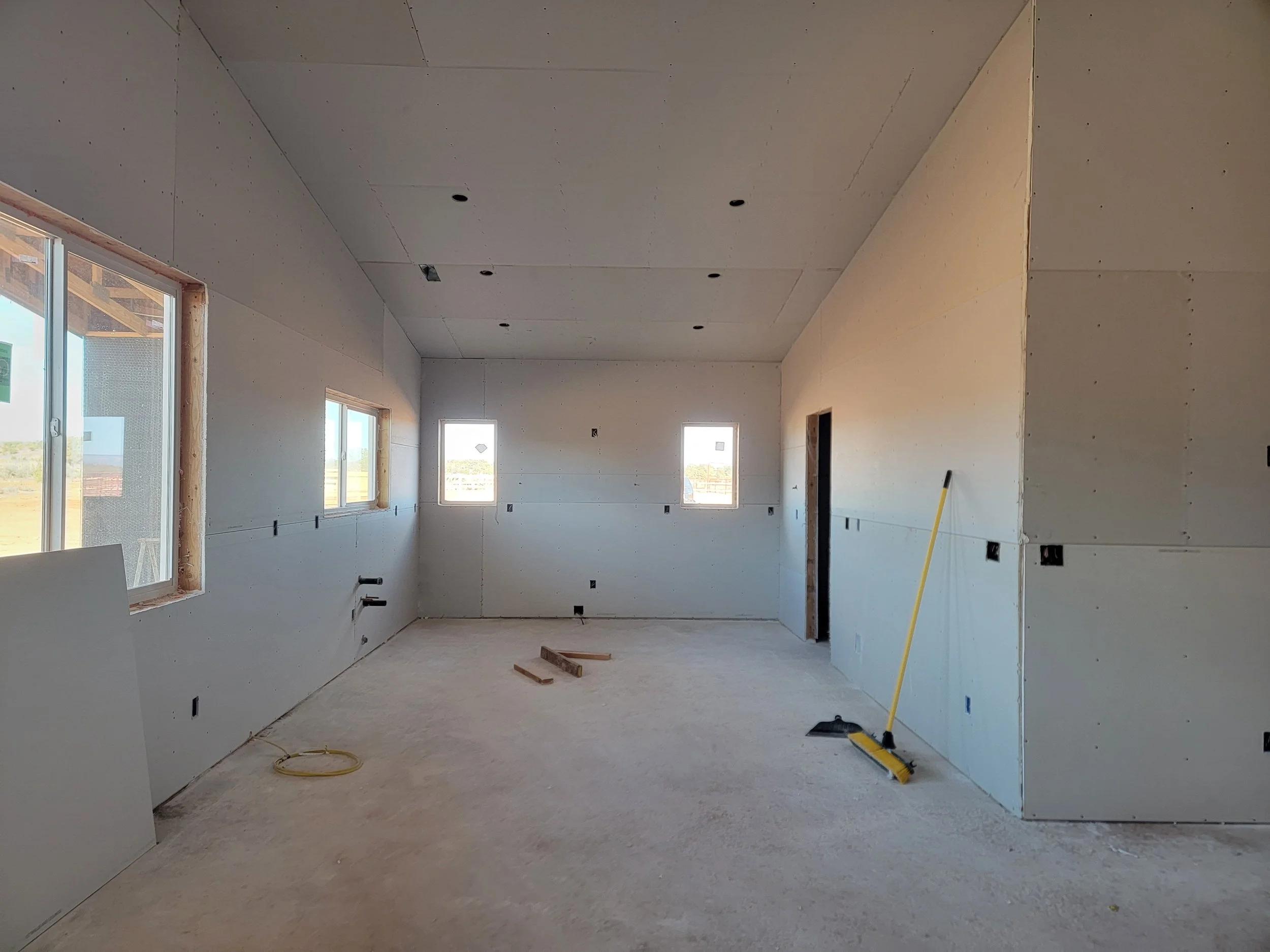 Interior of a house under construction with drywall walls and ceiling, multiple windows, construction tools on the floor, and an open doorway.