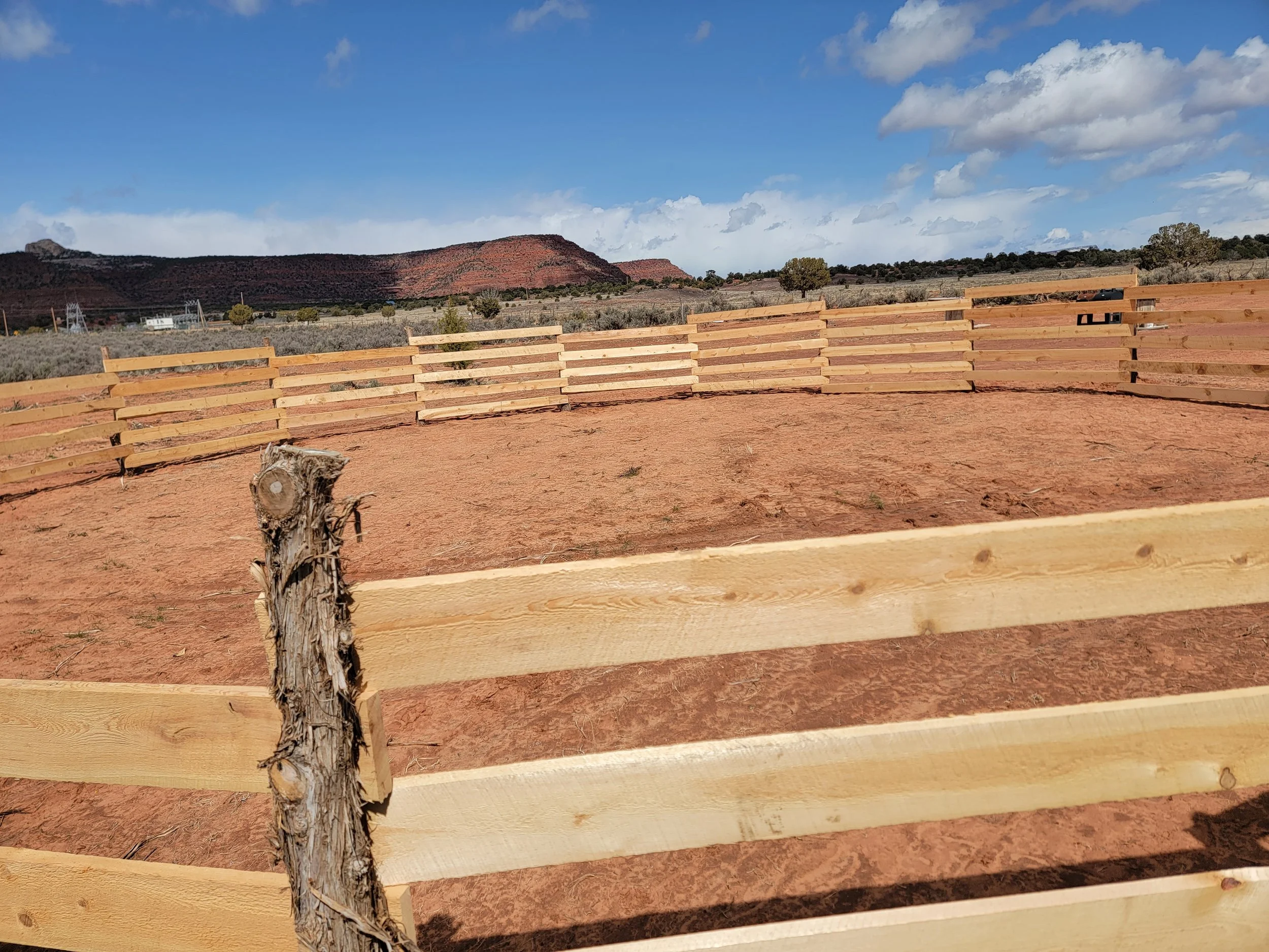 Wooden fence with red dirt ground in a rural landscape, hills in the background, and a partly cloudy sky.