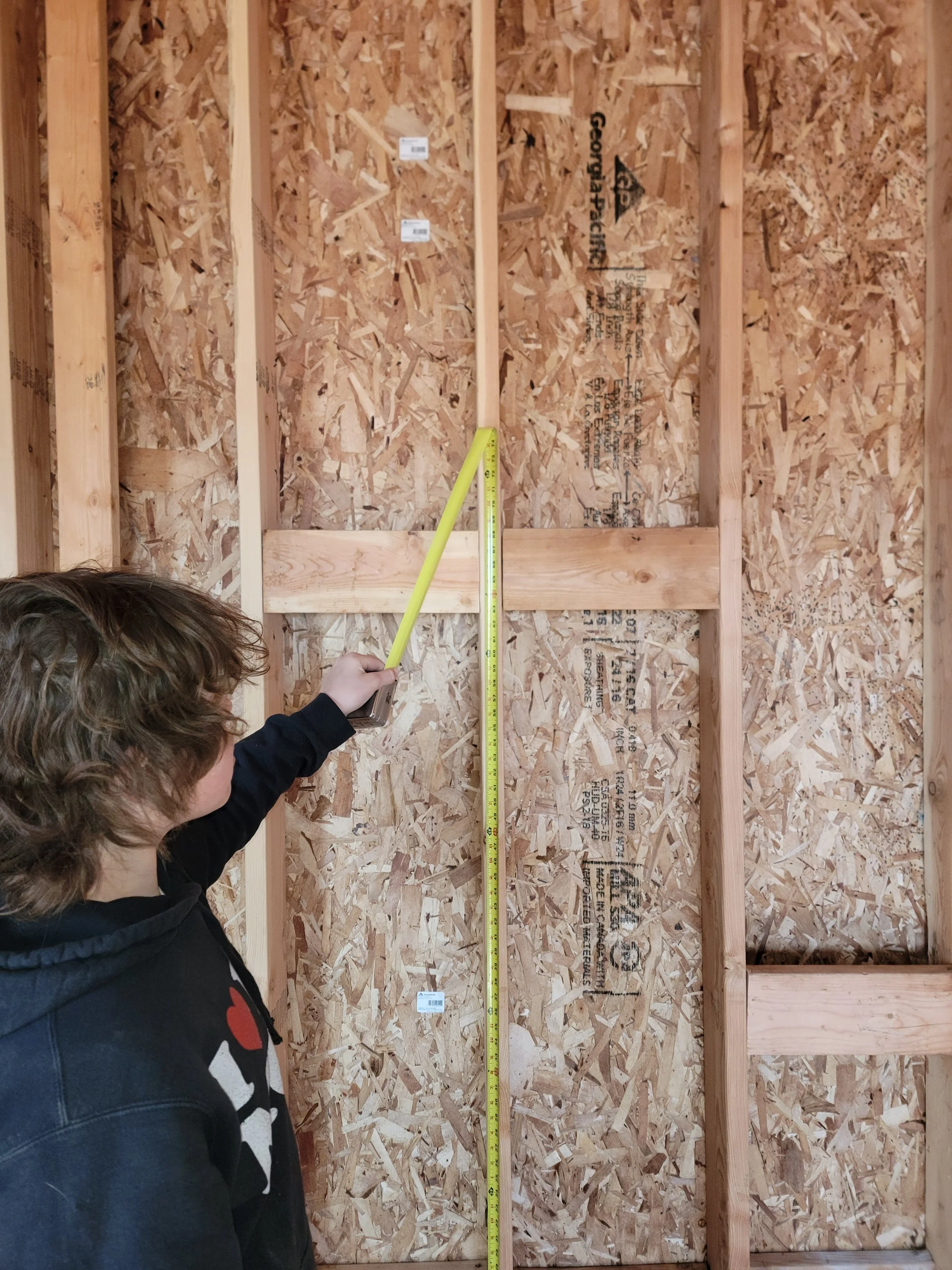 A person in a black hoodie is measuring a wall frame with a yellow tape measure, inside an unfinished wooden structure with oriented strand board sheathing.