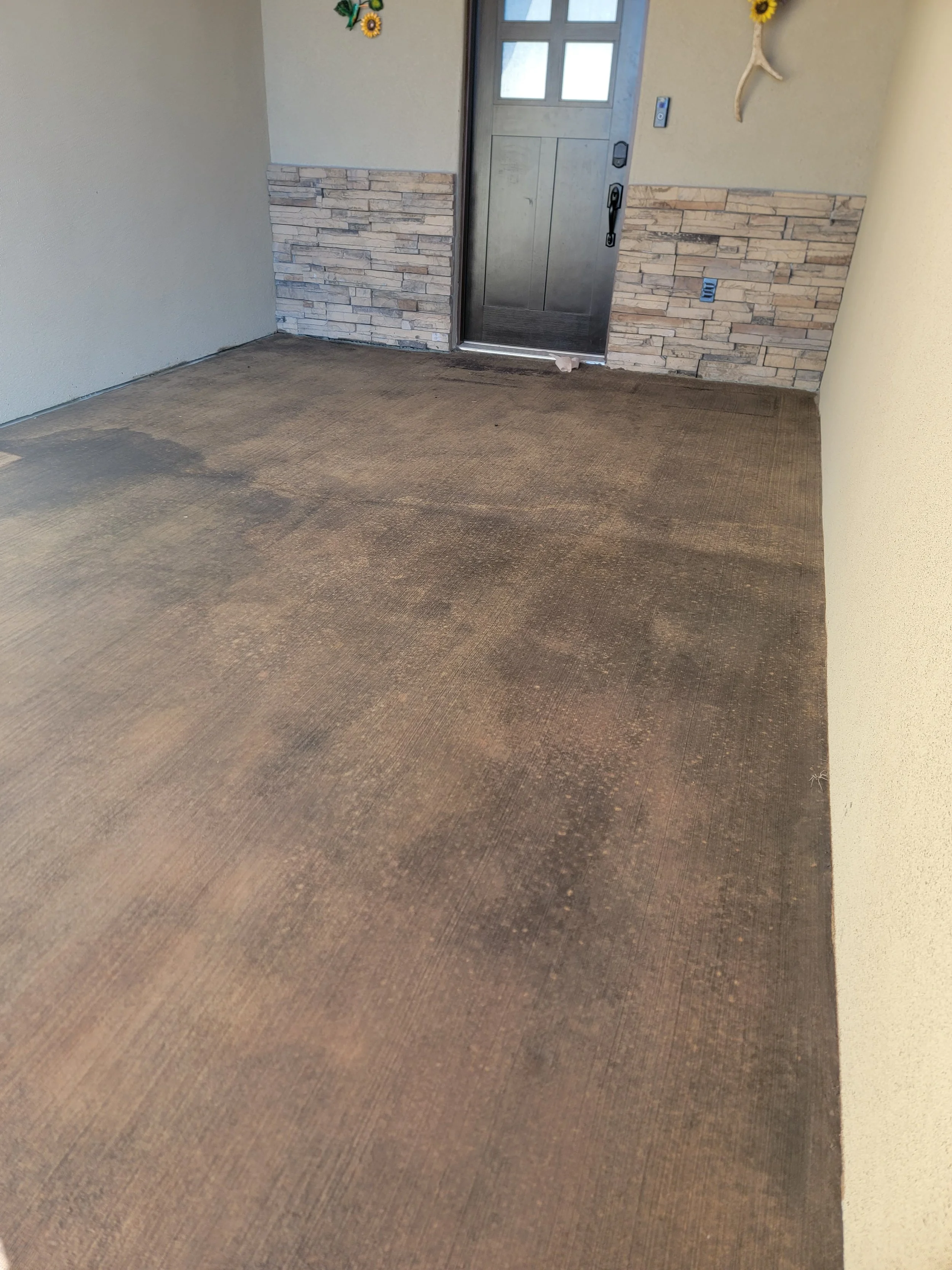 A covered porch with a brown textured floor, a gray door with four glass panels, and decorative sunflower wall accents.