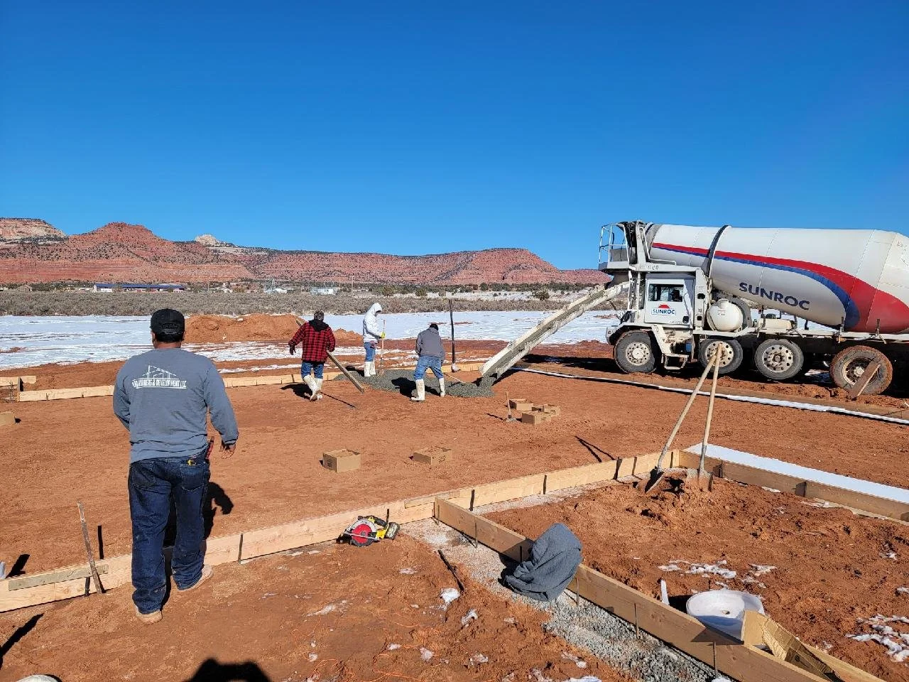 Construction site with workers pouring concrete from a concrete mixer truck on a clear, sunny day with a red rock landscape in the background.