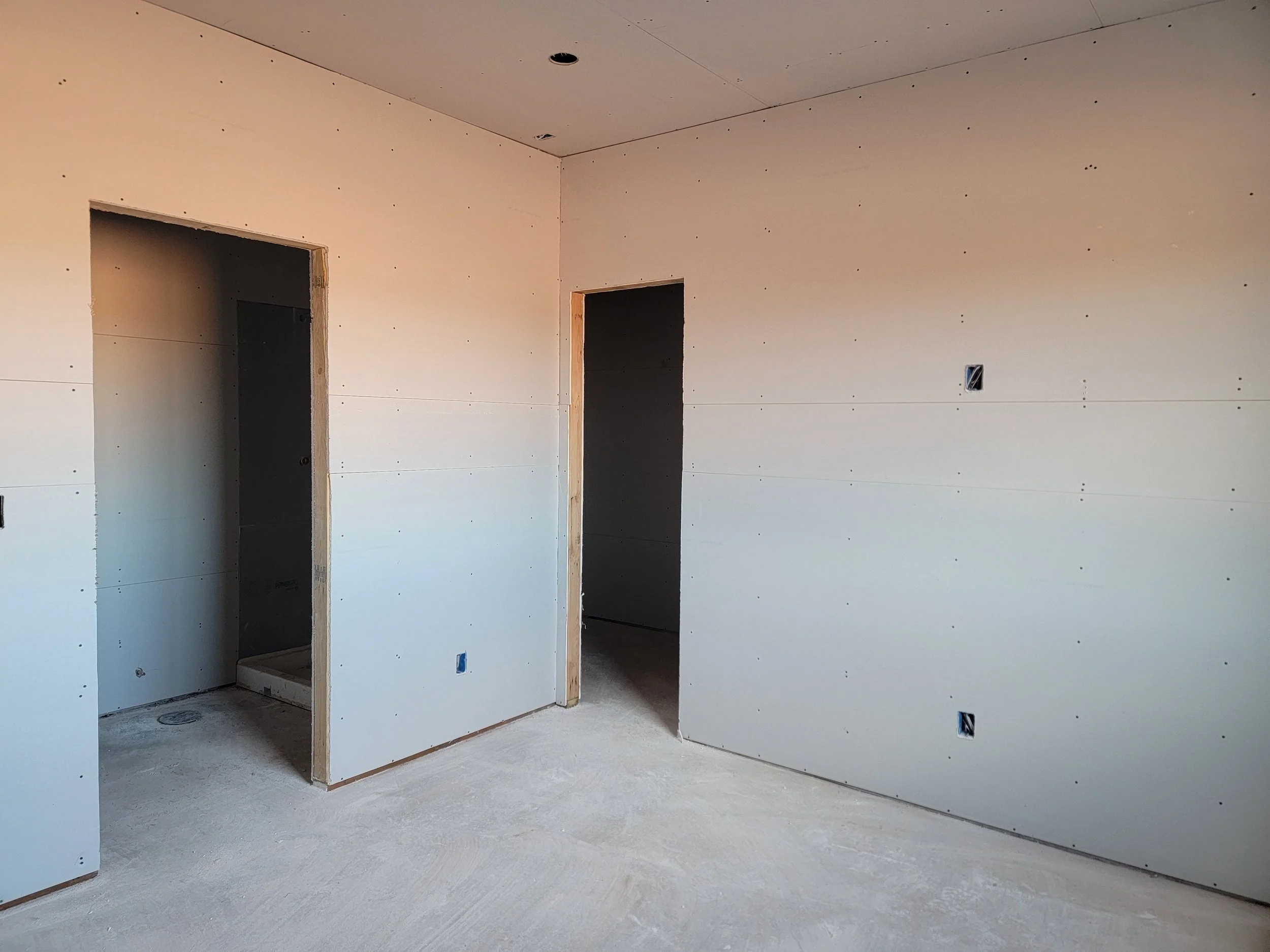 Interior of a house under construction, with unfinished drywall installed on walls and ceiling, and electrical outlets and switches not yet installed.