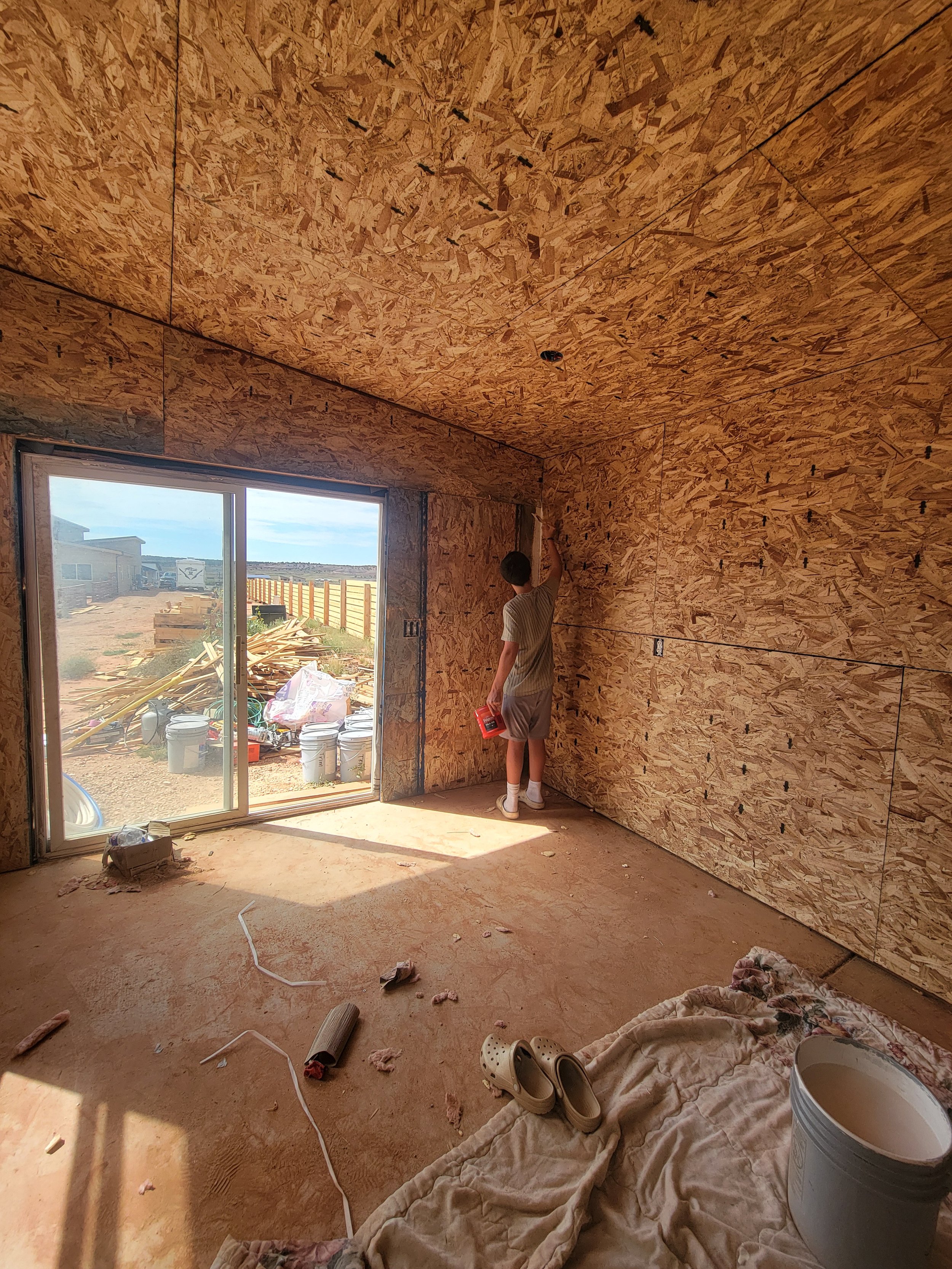 An unfinished room with plywood walls and ceiling, a sliding glass door leading outside, and various construction materials and tools scattered on the floor, including a bucket, cloth, shoes, and debris. A person stands near the corner, reaching up t
