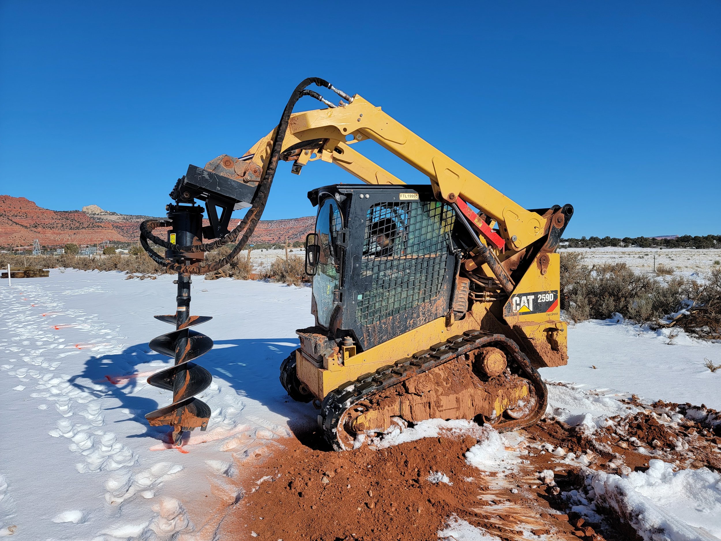 A yellow Caterpillar backhoe with a large auger attachment, situated on a snowy landscape with footprints and a dirt patch, under a clear blue sky.