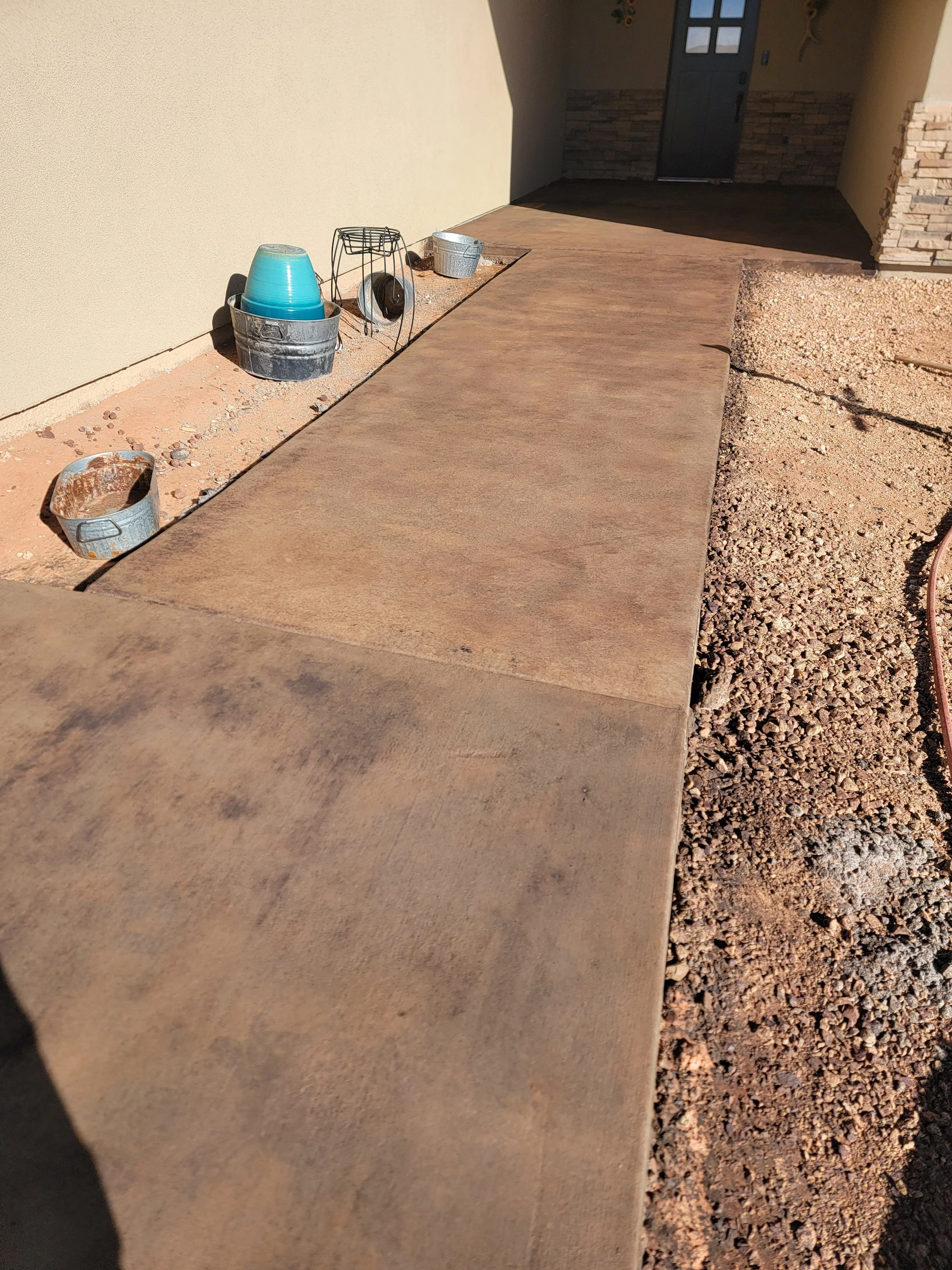 Construction site with a concrete walkway, construction buckets, a dish drainer with dishes, and gravel next to a beige wall and a dark door in the background.